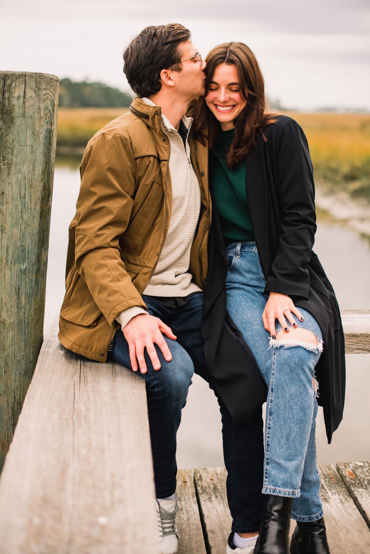 A couple sits together on a wooden pier by a lake wearing casual fall clothing and sharing intimate moments.