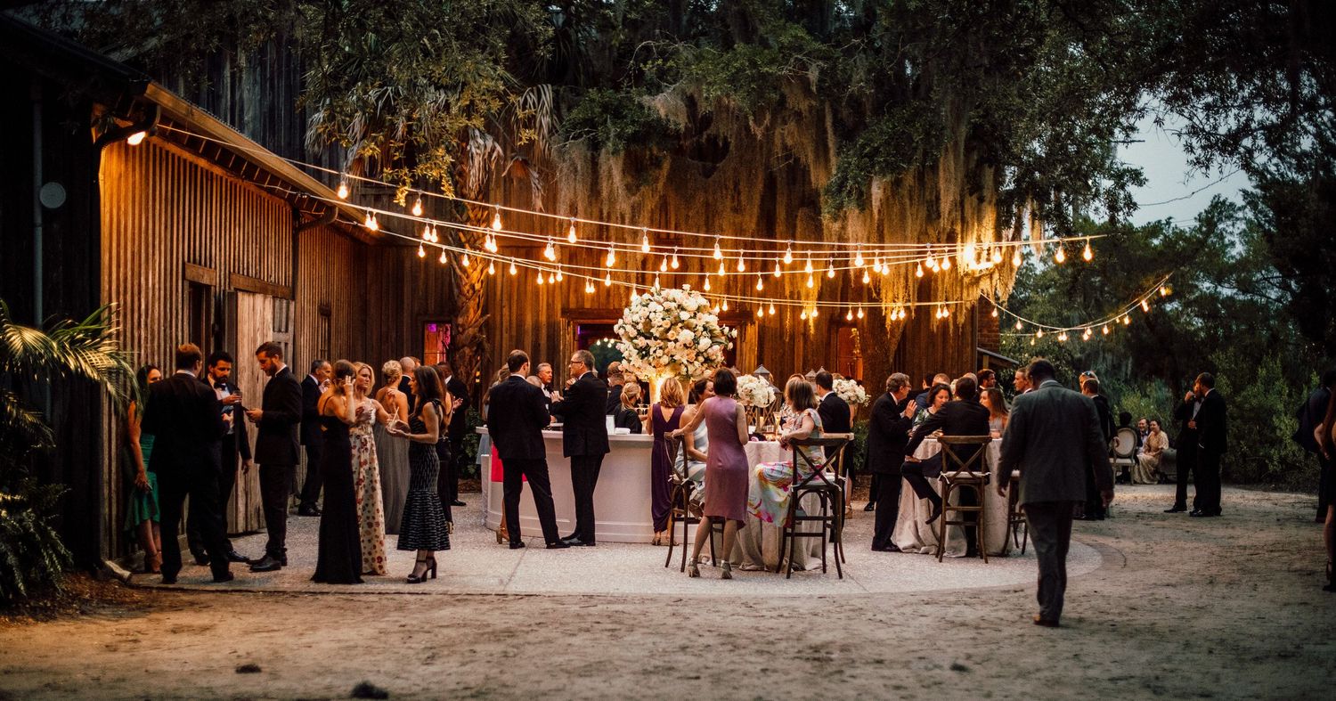 String lights illuminate an outdoor evening wedding reception with guests mingling under a warm glow at dusk.