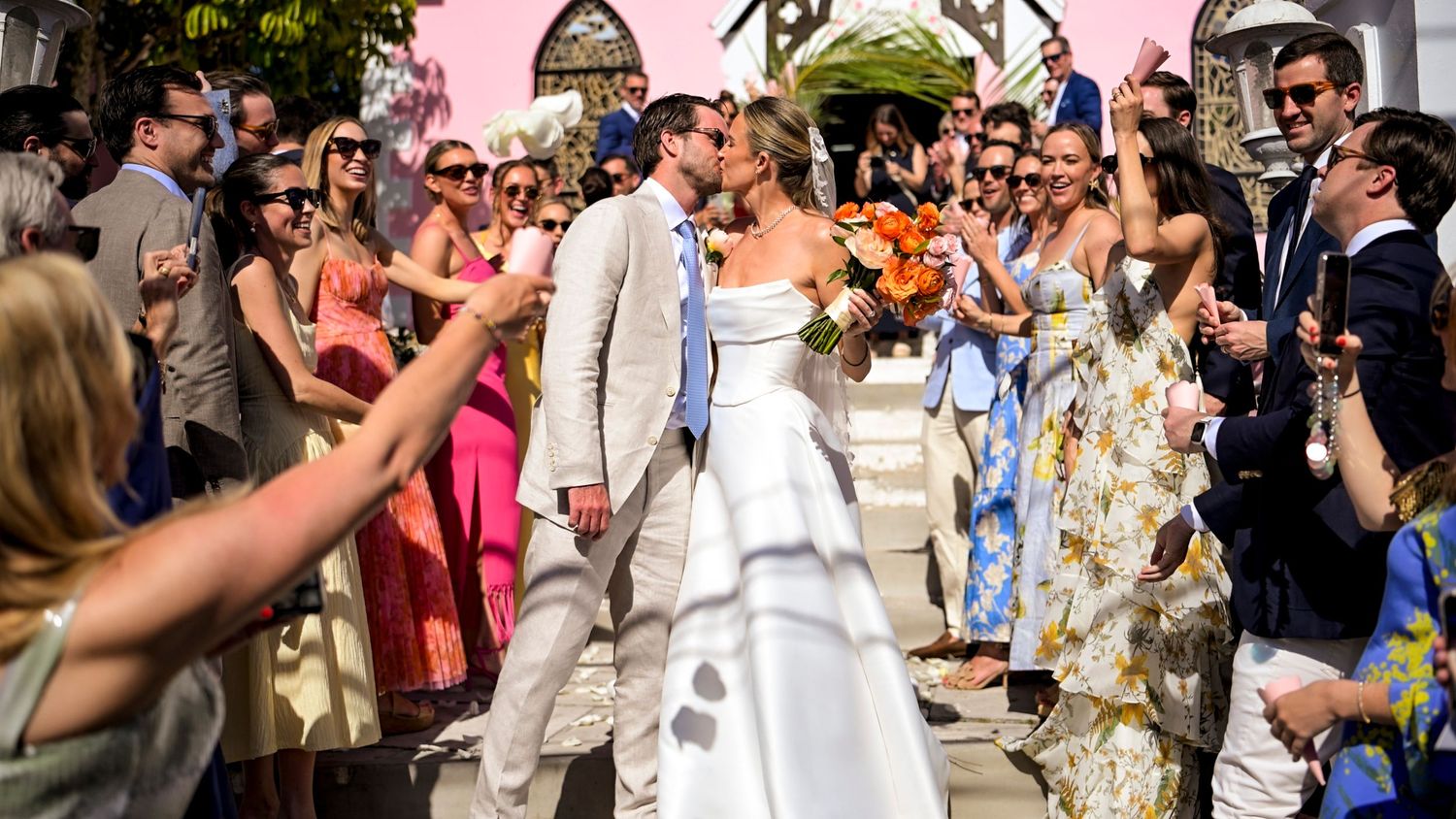 Wedding guests celebrate and take photos as the newlyweds exit through a pink-walled courtyard.