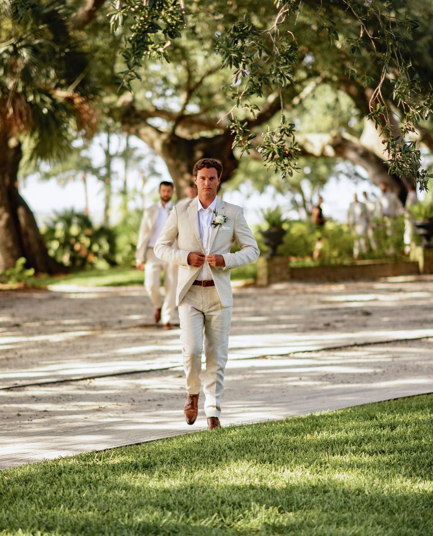 Groomsmen in tan suits run playfully across a grassy lawn under tree branches at an outdoor wedding venue.