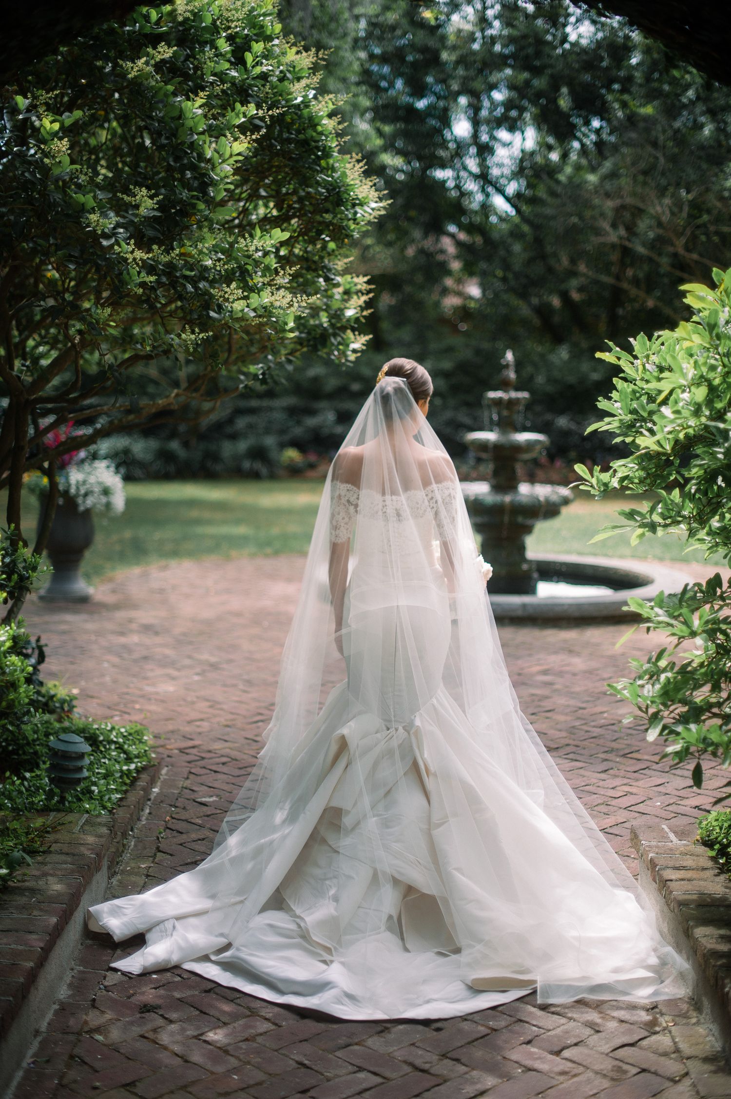 A bride in a flowing white wedding gown and veil sits gracefully on a brick path surrounded by lush garden greenery.