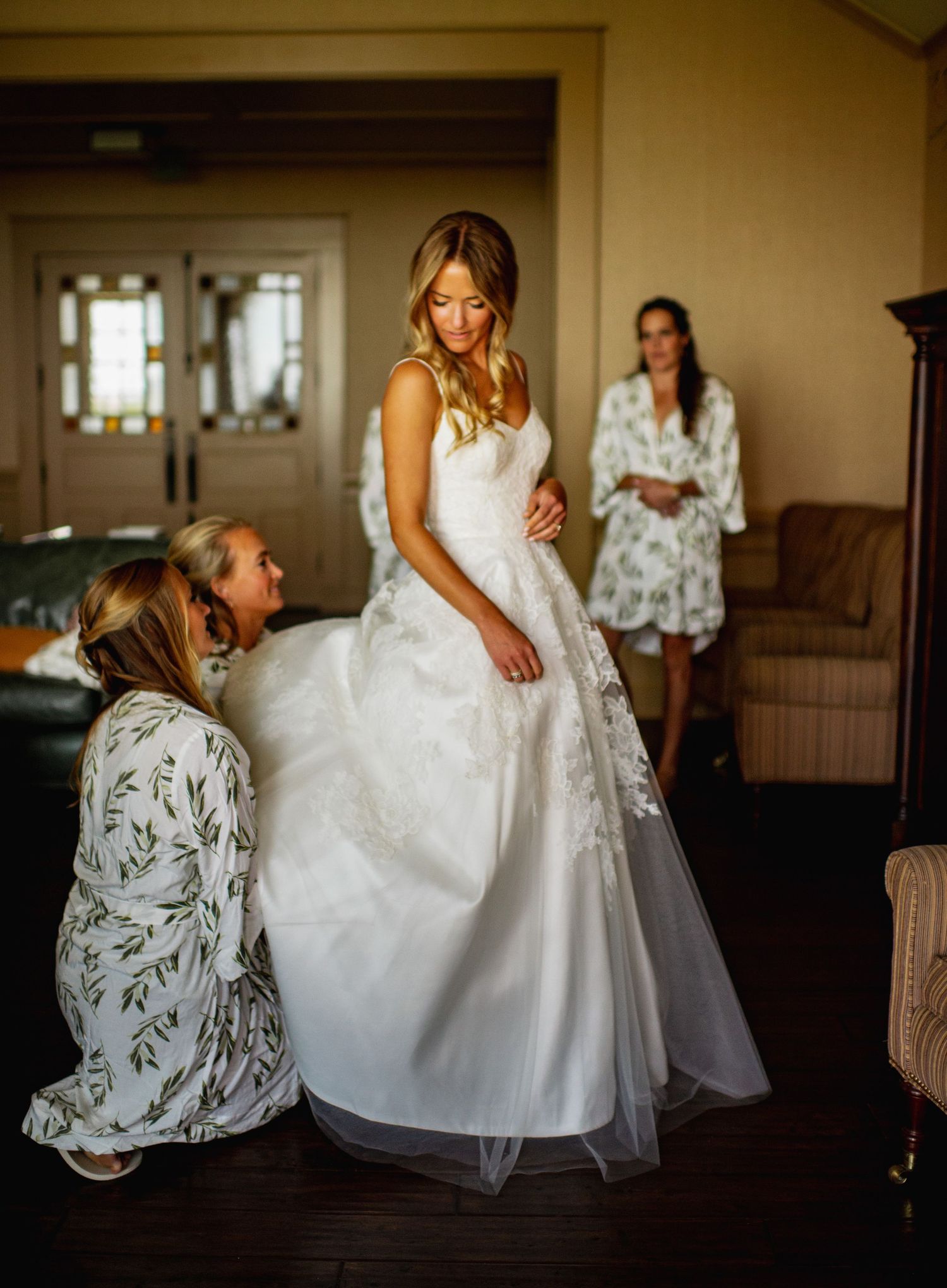 A bride in a flowing white wedding gown gets ready with bridesmaids in floral robes in a sunlit room.
