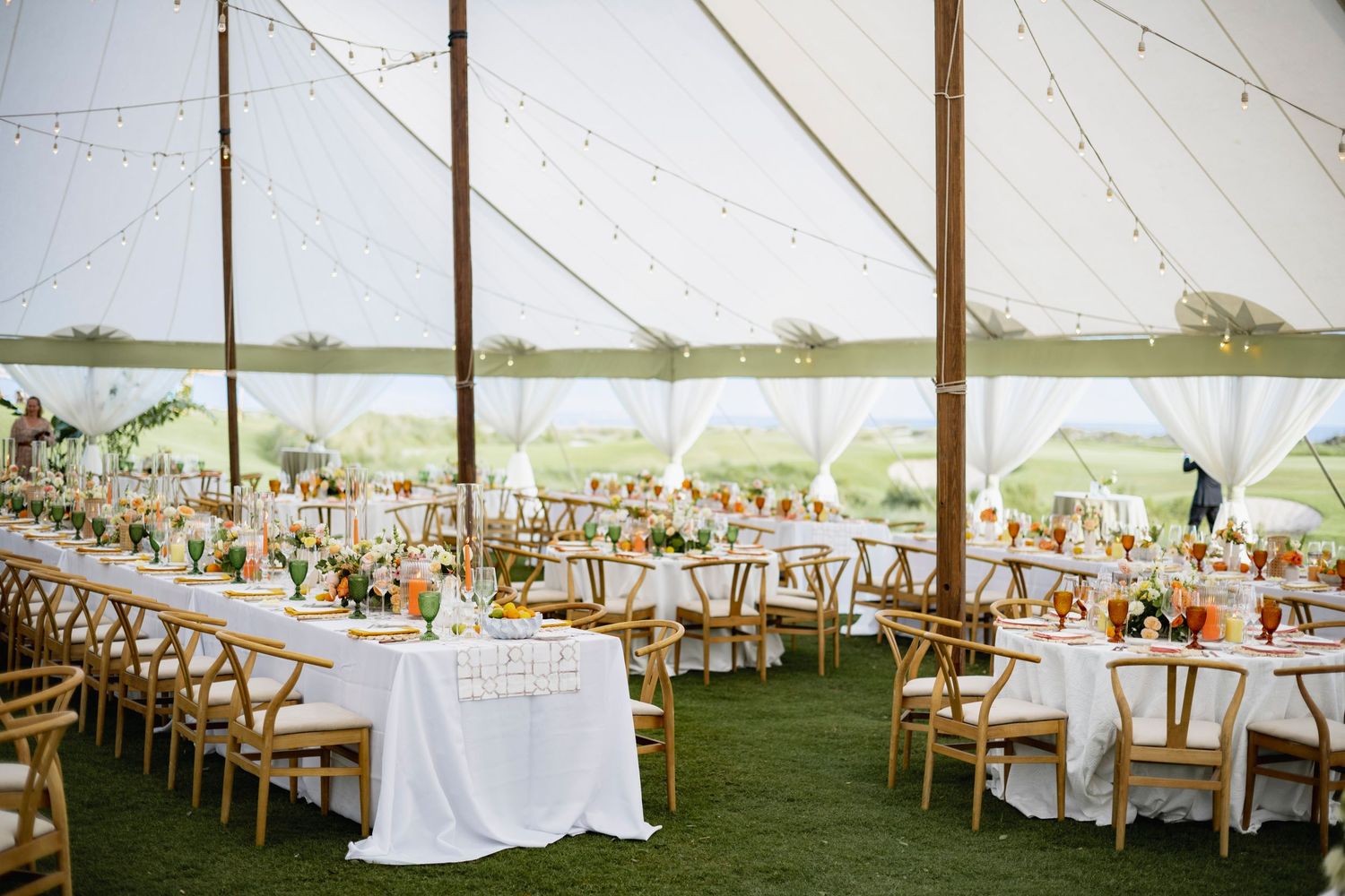 Elegant outdoor wedding reception setup with white tables and wooden chairs under a tent overlooking a scenic landscape.