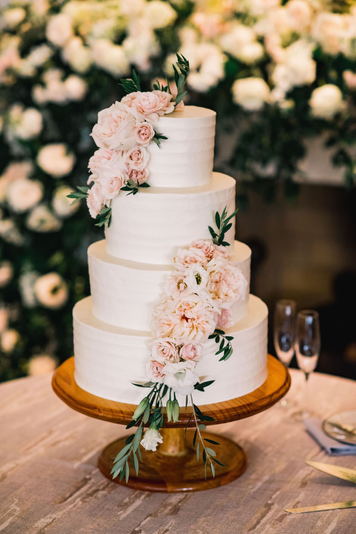 Elegant three-tiered white wedding cake decorated with soft pink flowers and greenery on a wooden cake stand.