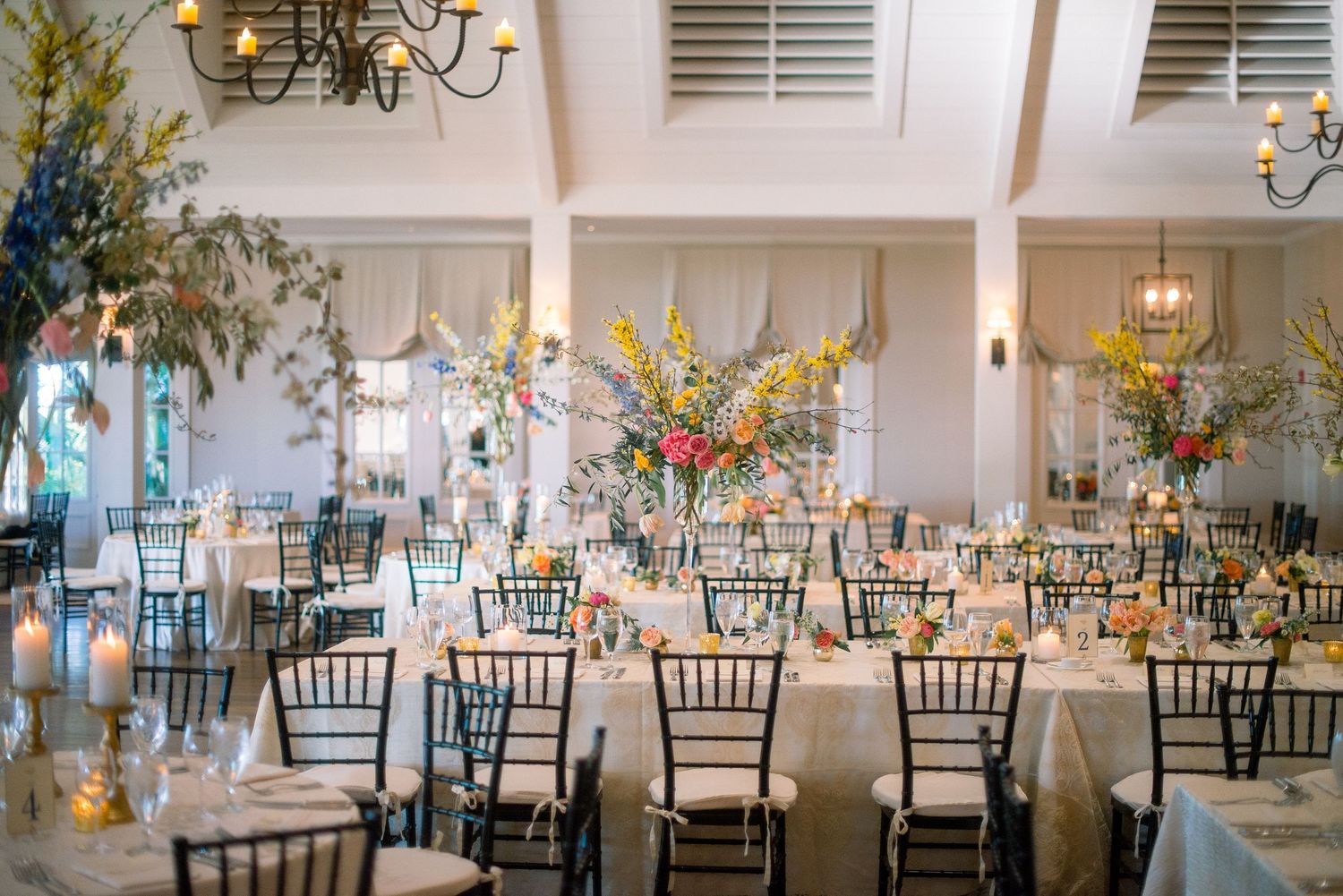 Elegant wedding reception venue with tall floral centerpieces, white tablecloths and dark chiavari chairs in a bright ballroom.