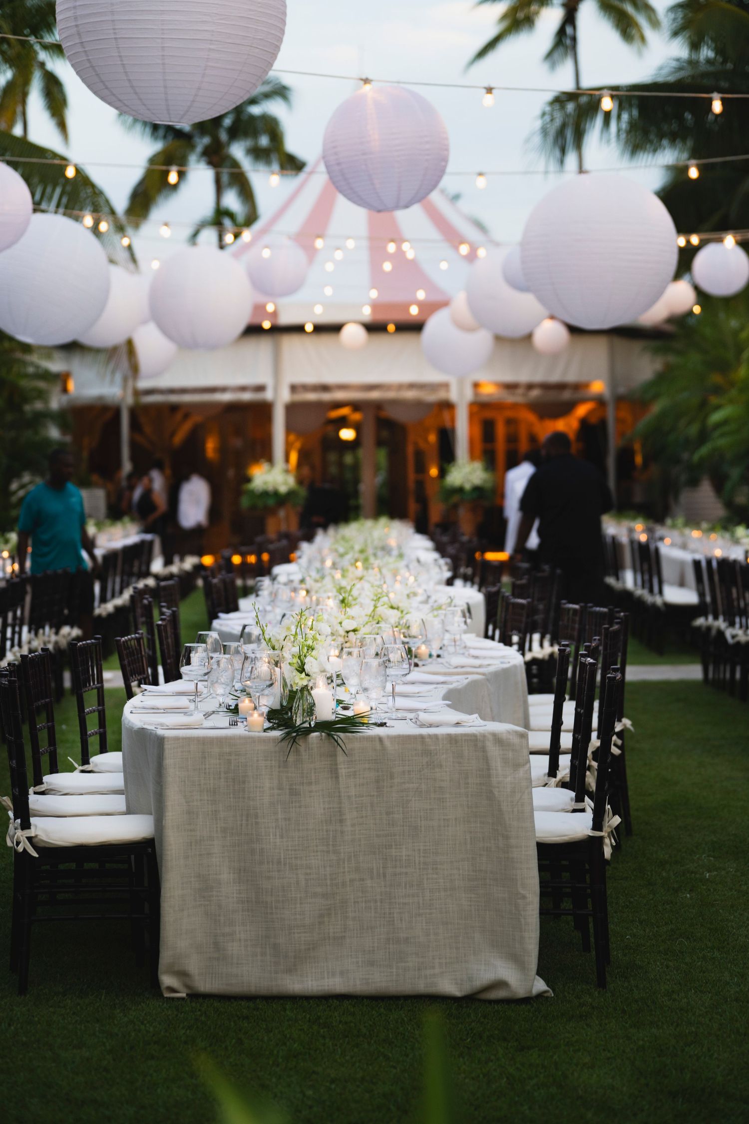 Elegant outdoor wedding reception setup with white paper lanterns and long tables decorated in neutral linens and florals.