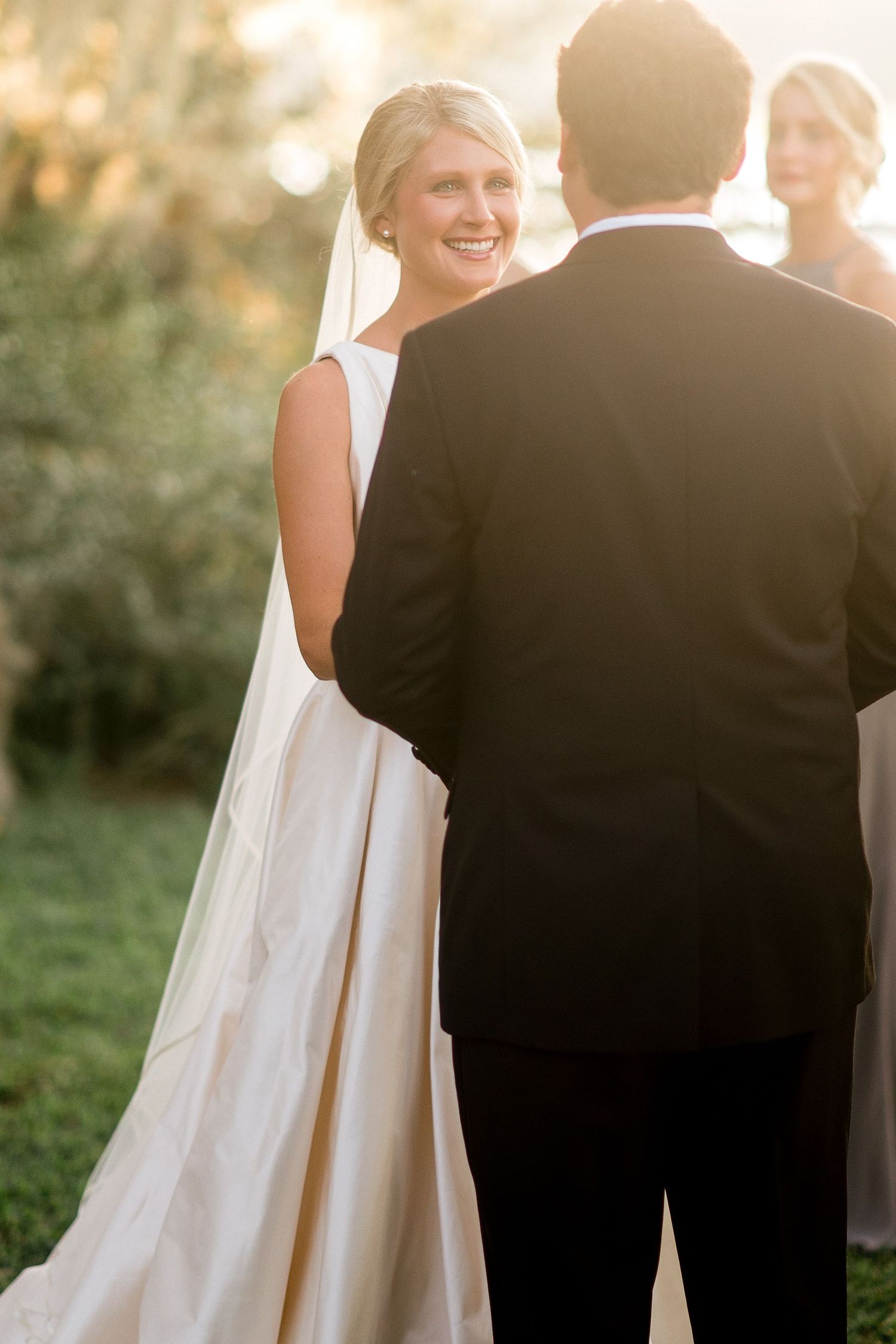 A bride in a flowing white dress stands with her groom in an outdoor garden setting during golden hour.