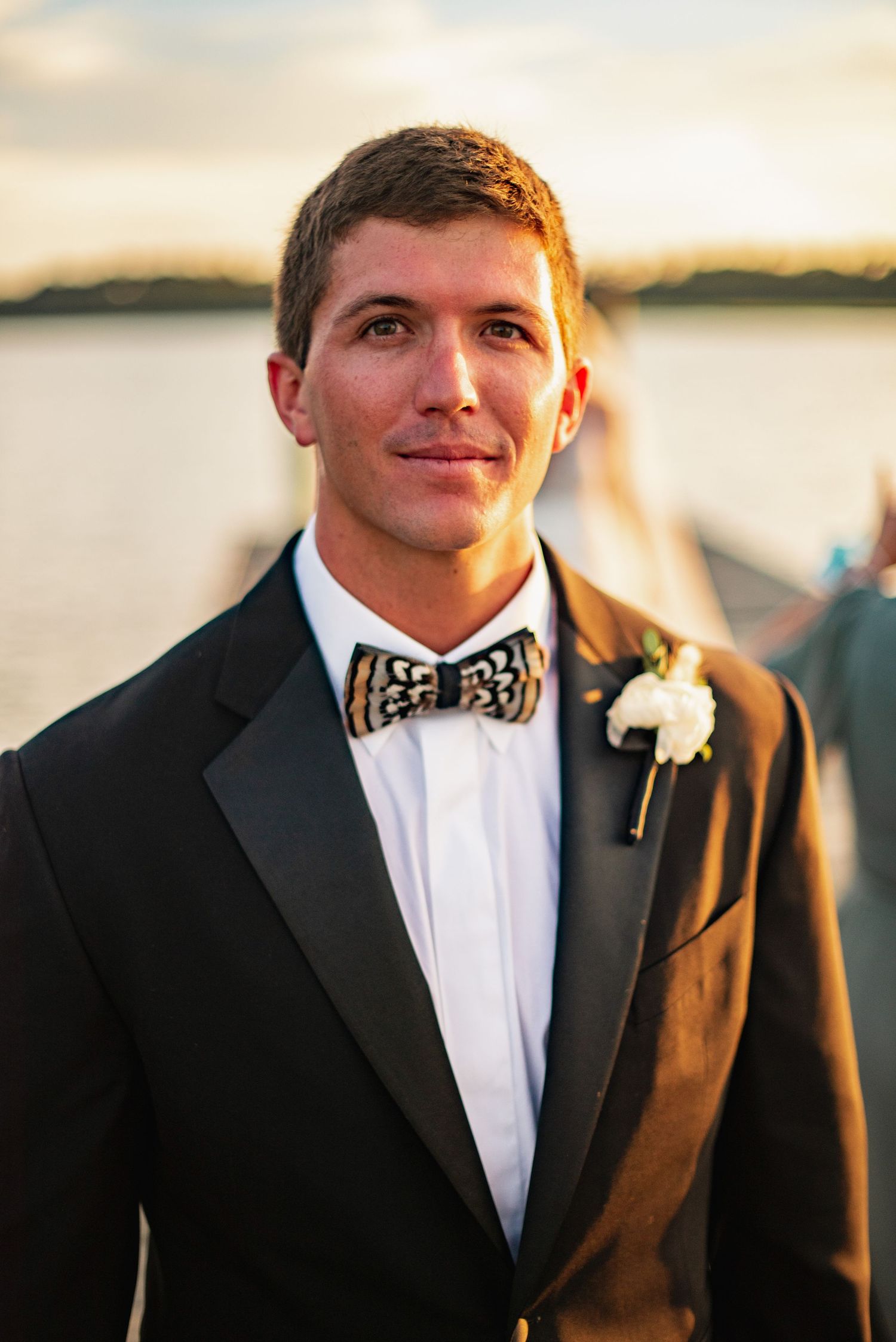 A groom in a black tuxedo and polka dot bowtie smiles at sunset during a waterfront wedding ceremony.