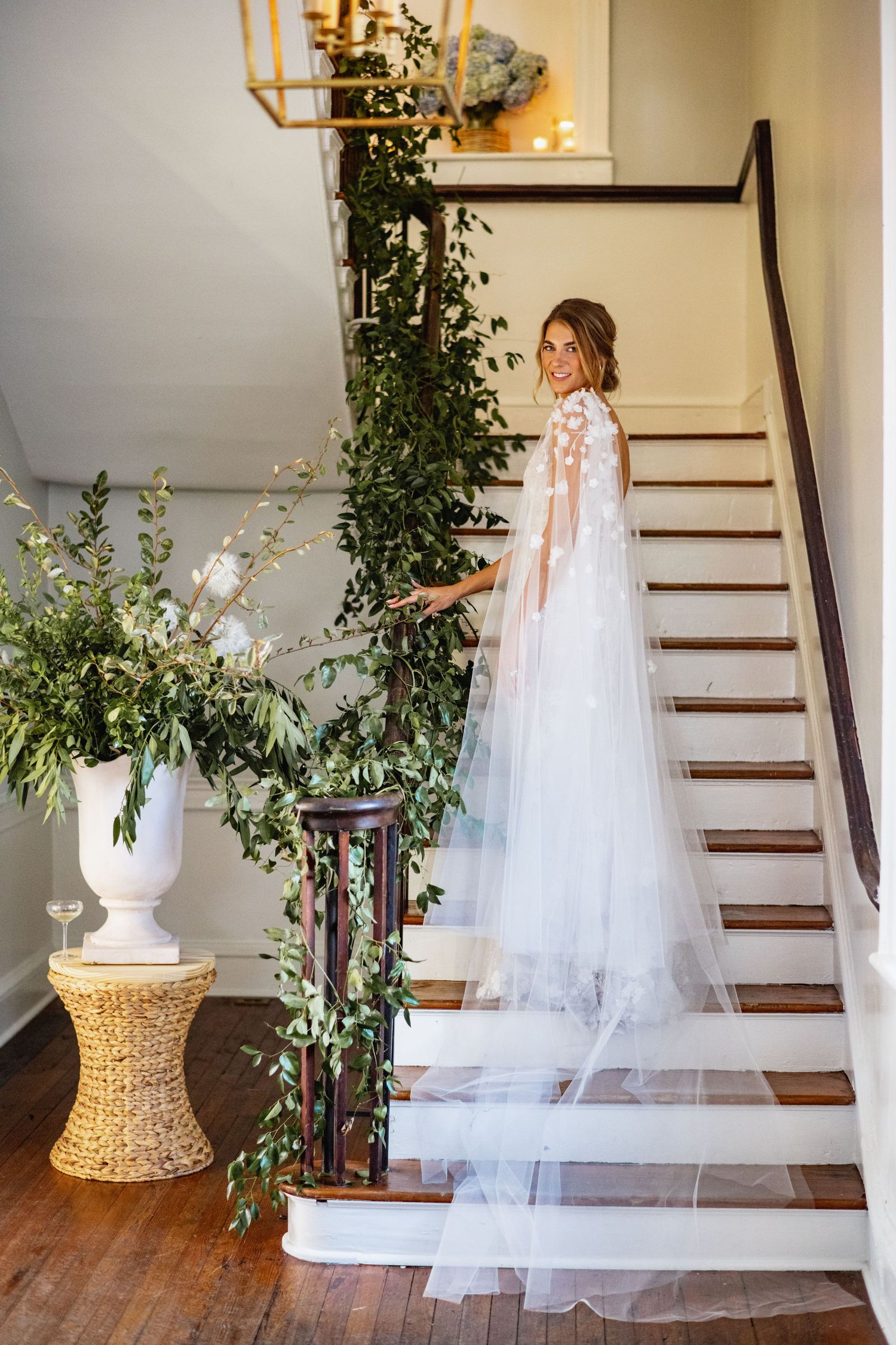 A flowing white wedding dress and veil displayed on a wooden staircase with greenery and floral decorations.