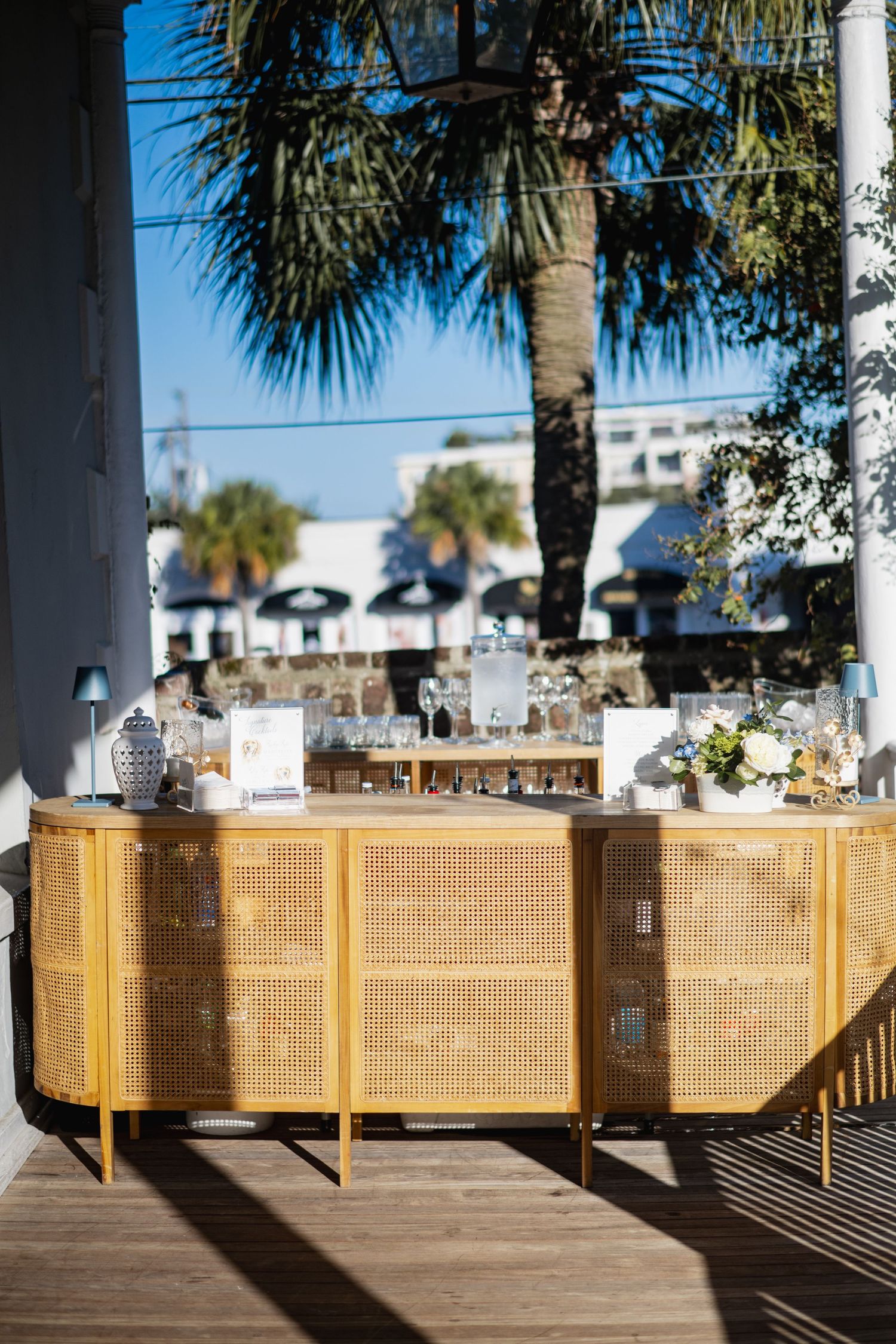 Wooden deck railing with palm trees and beach buildings visible in the background on a sunny day.