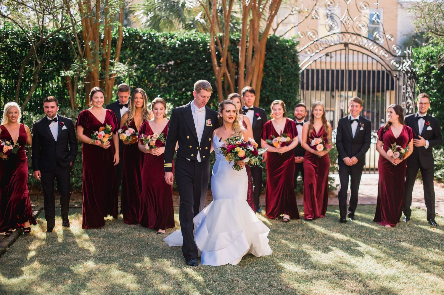 Wedding party in burgundy dresses and black suits poses together outdoors in front of trees and greenery.