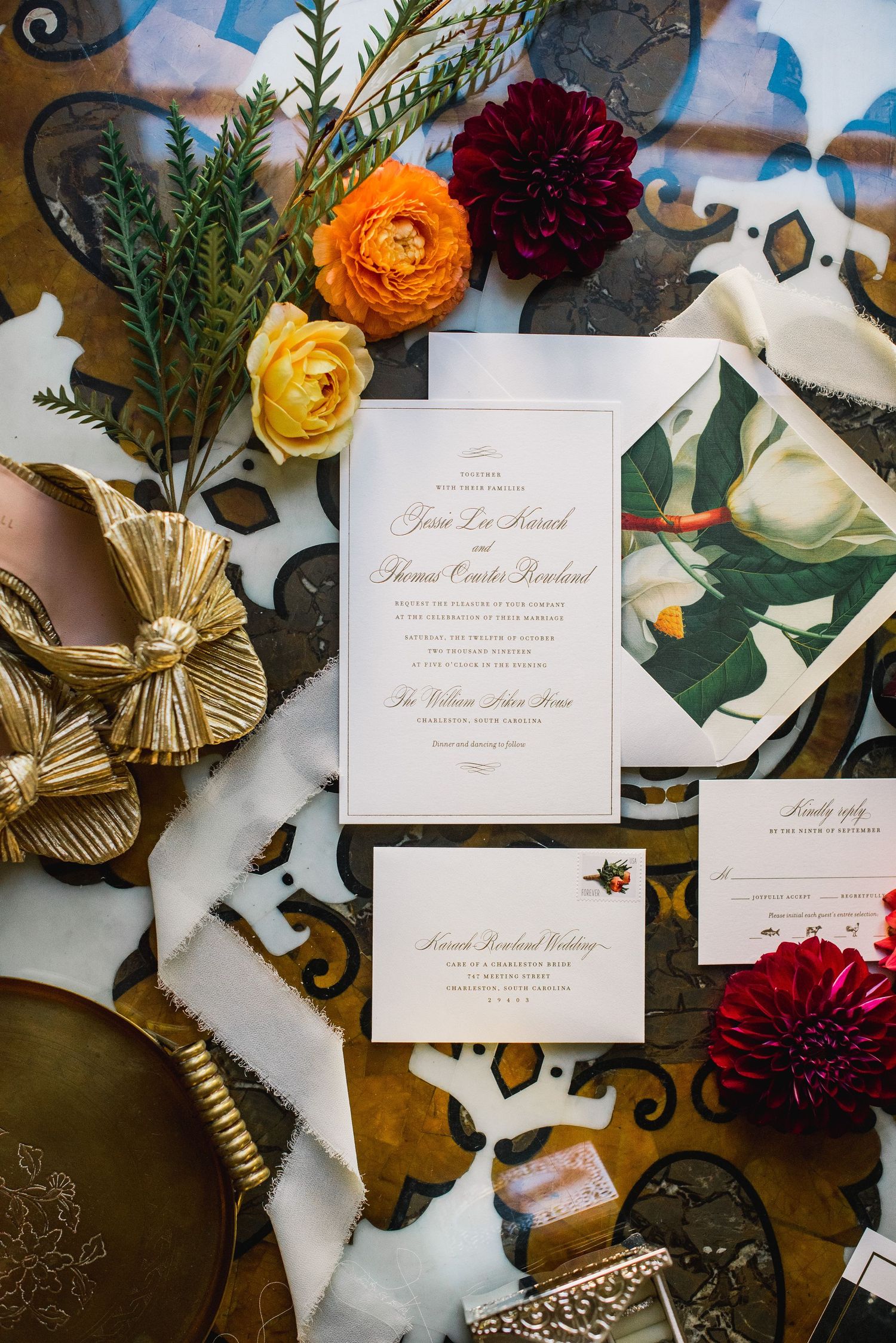 Elegant wedding invitation suite with gold accents, burgundy dahlias, and tropical greenery arranged on a textured surface.