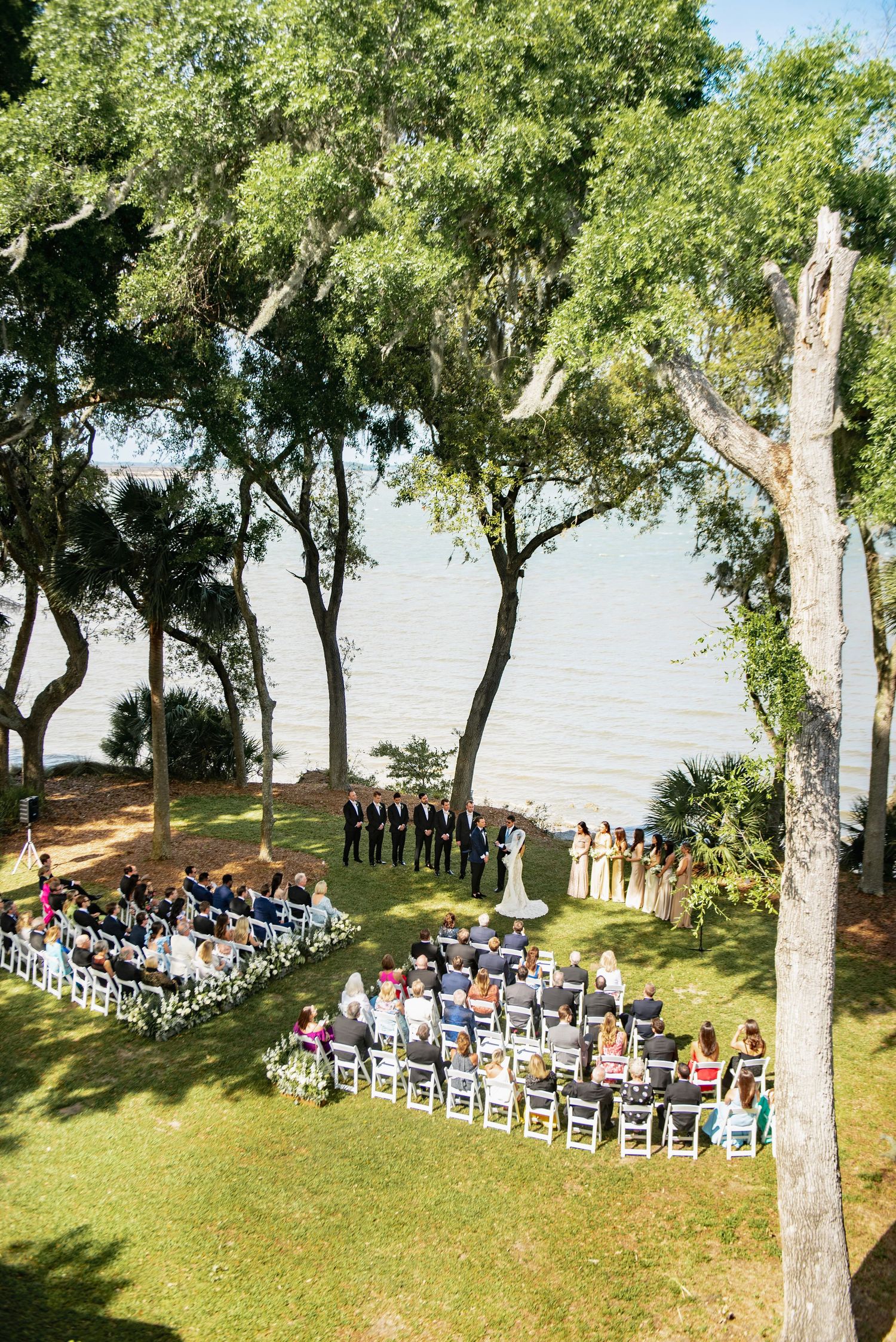Outdoor wedding ceremony setup with white chairs arranged in curved rows under shady trees by the waterfront.