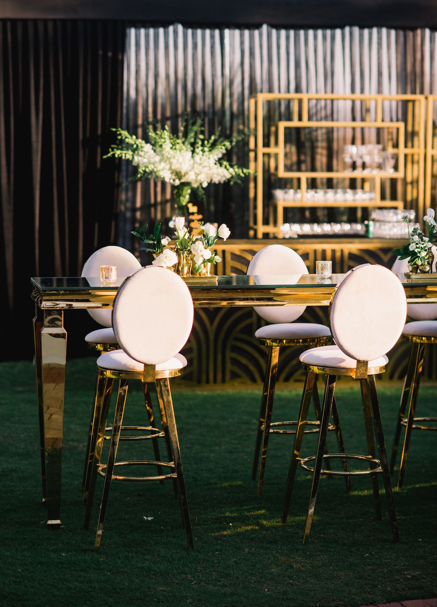 Elegant wedding reception setup with white oval chairs and gold accents against green carpet and bamboo backdrop.