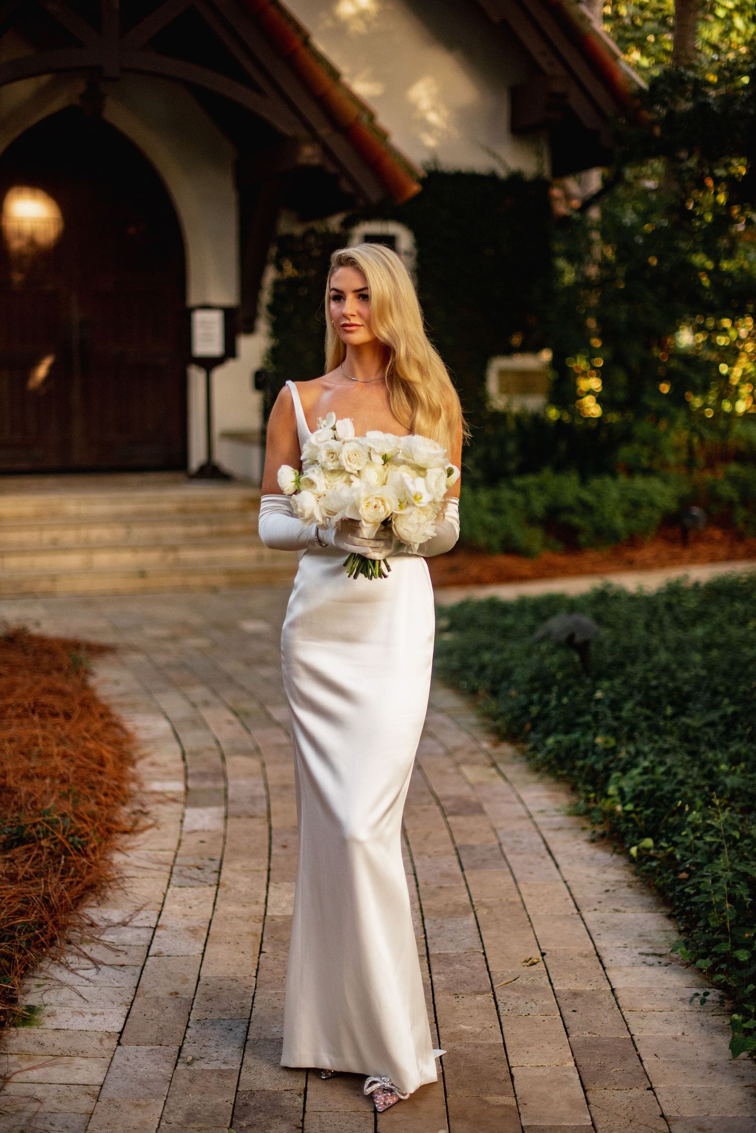 A bride in an elegant white off-shoulder wedding gown walks along a brick pathway holding a white floral bouquet.