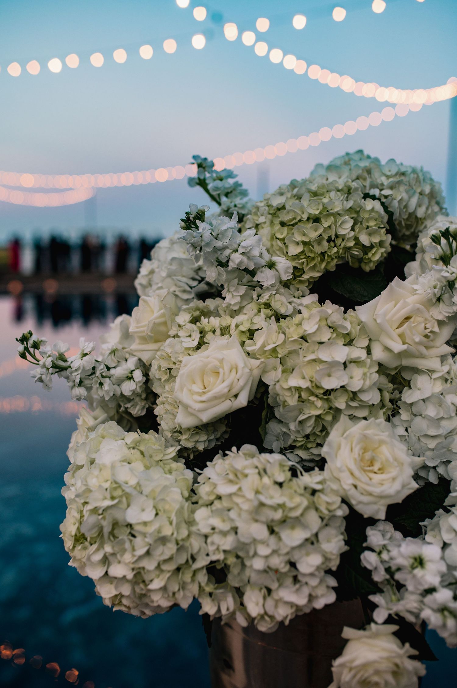 White roses and hydrangeas create an elegant floral arrangement against a sunset sky with twinkling string lights.
