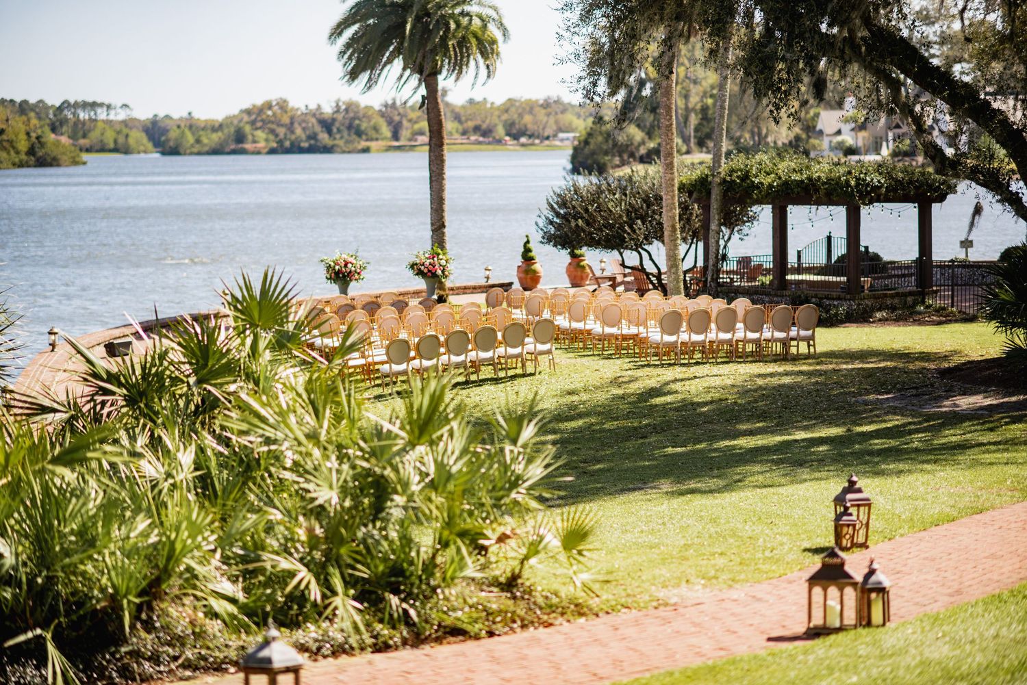 Palm trees and chairs line a waterfront ceremony setup with a picturesque lake view in the background.