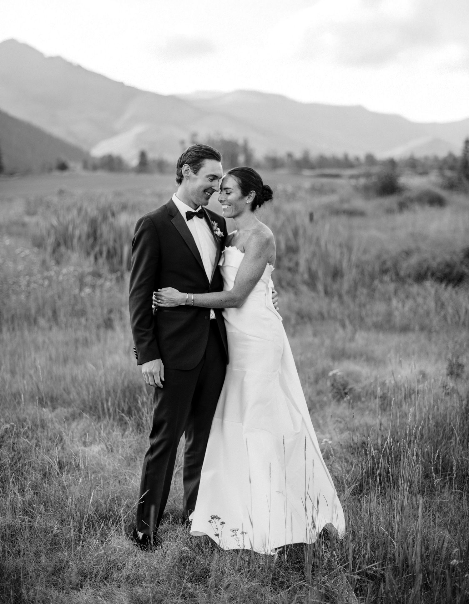 A romantic black and white wedding photo of a couple embracing in a meadow with mountains in the background.