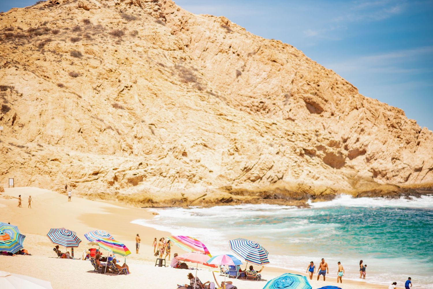 Colorful beach umbrellas dot a sandy beach beneath dramatic golden cliffs along the Baja coast in Cabo San Lucas