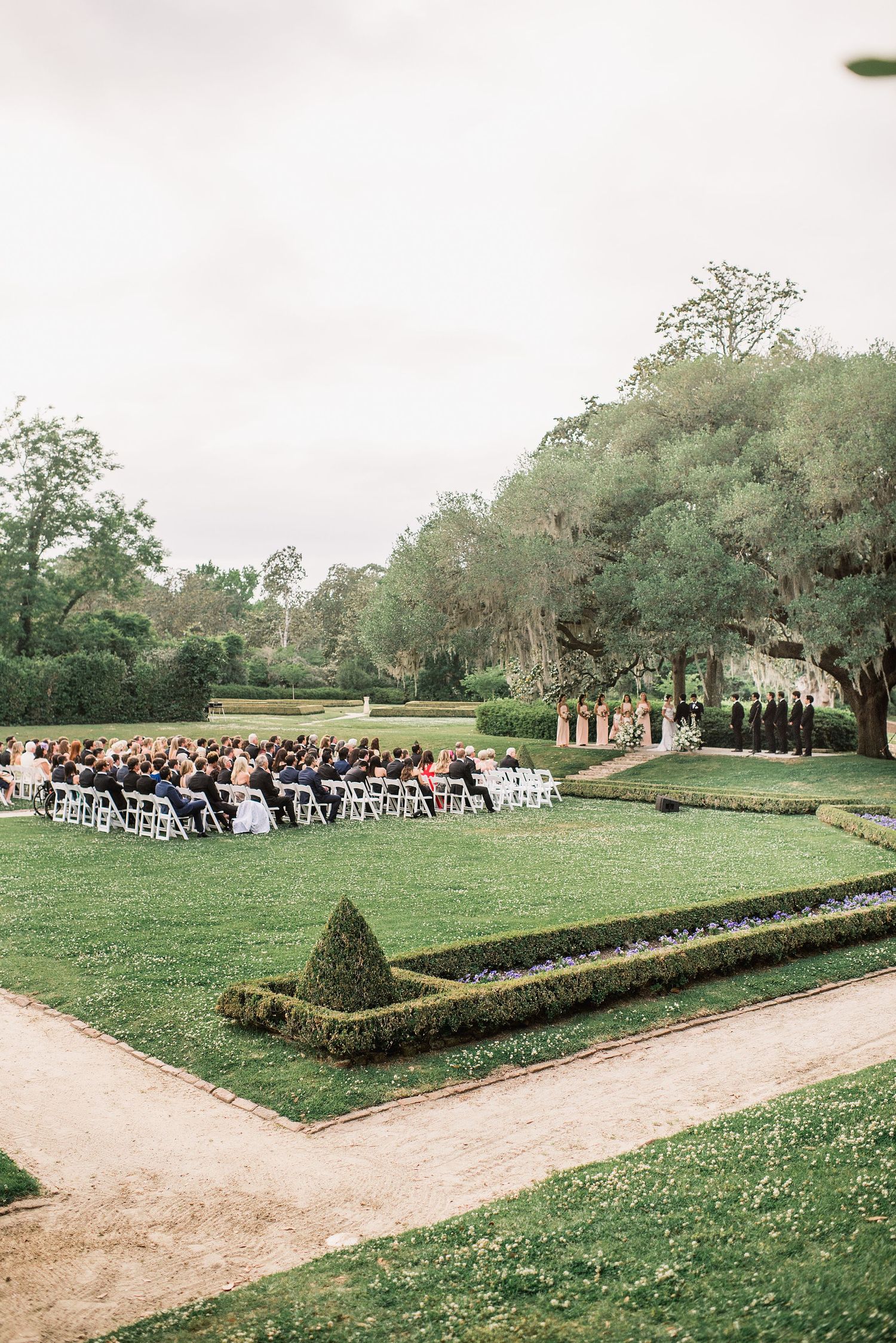 A panoramic view of an outdoor wedding ceremony setup with guests seated on white chairs in a manicured garden setting.