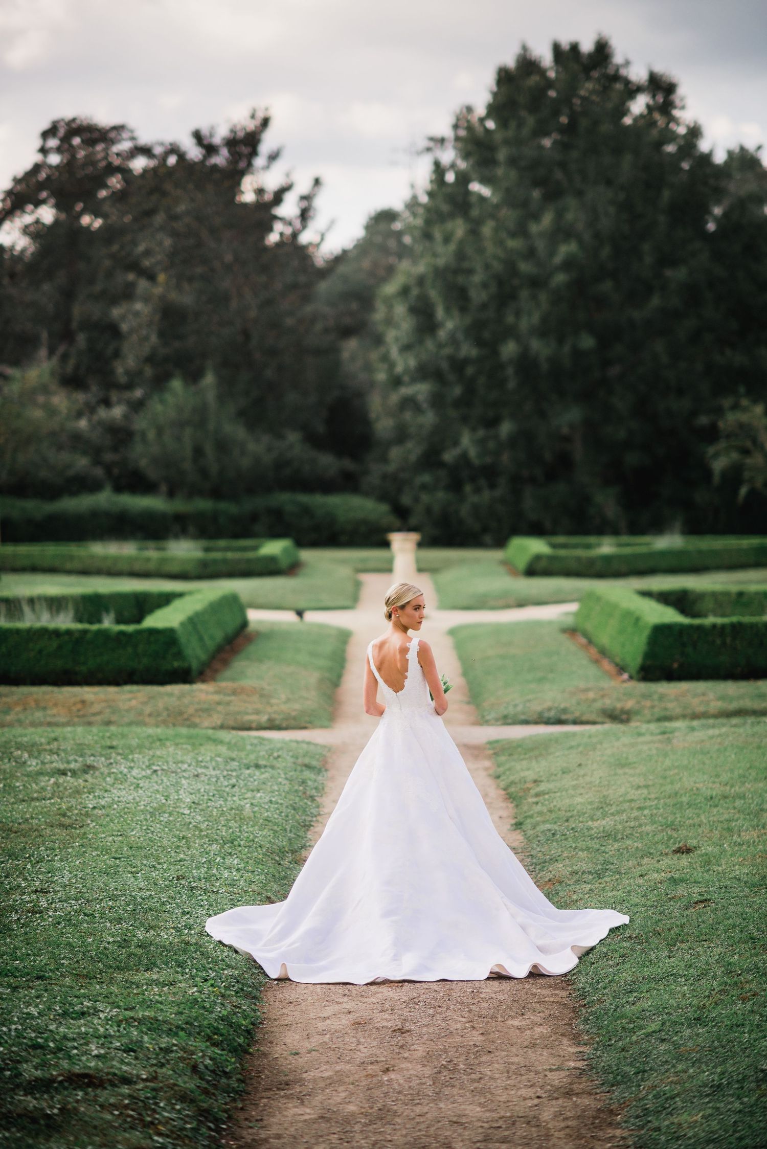 A person in a flowing white wedding gown walks along a garden path surrounded by manicured hedges.