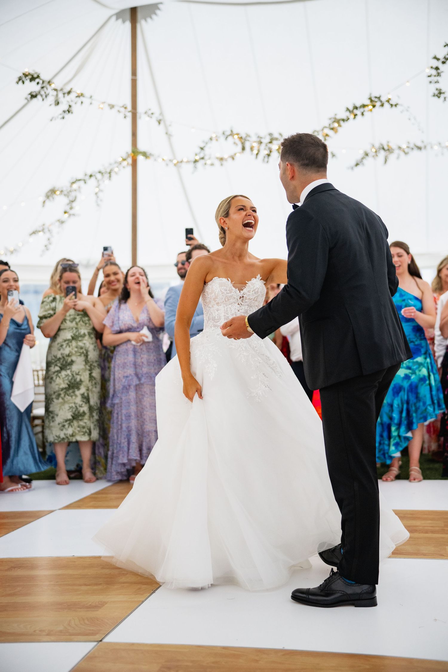Wedding couple dancing together under a white tent while guests cheer and take photos.