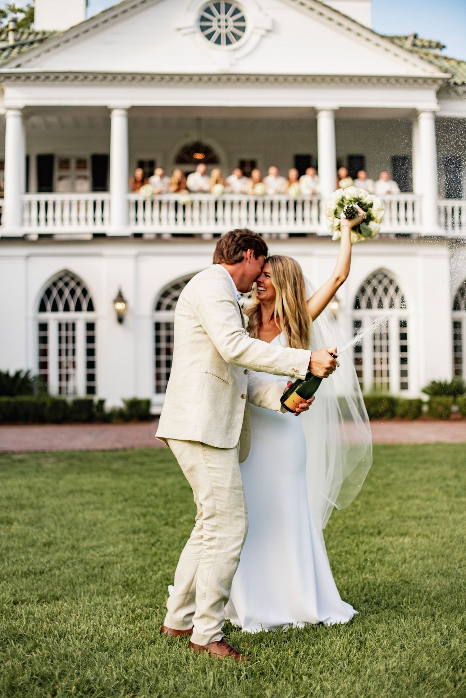 A couple in white wedding attire shares a romantic dip pose on a manicured lawn in front of an elegant white mansion.
