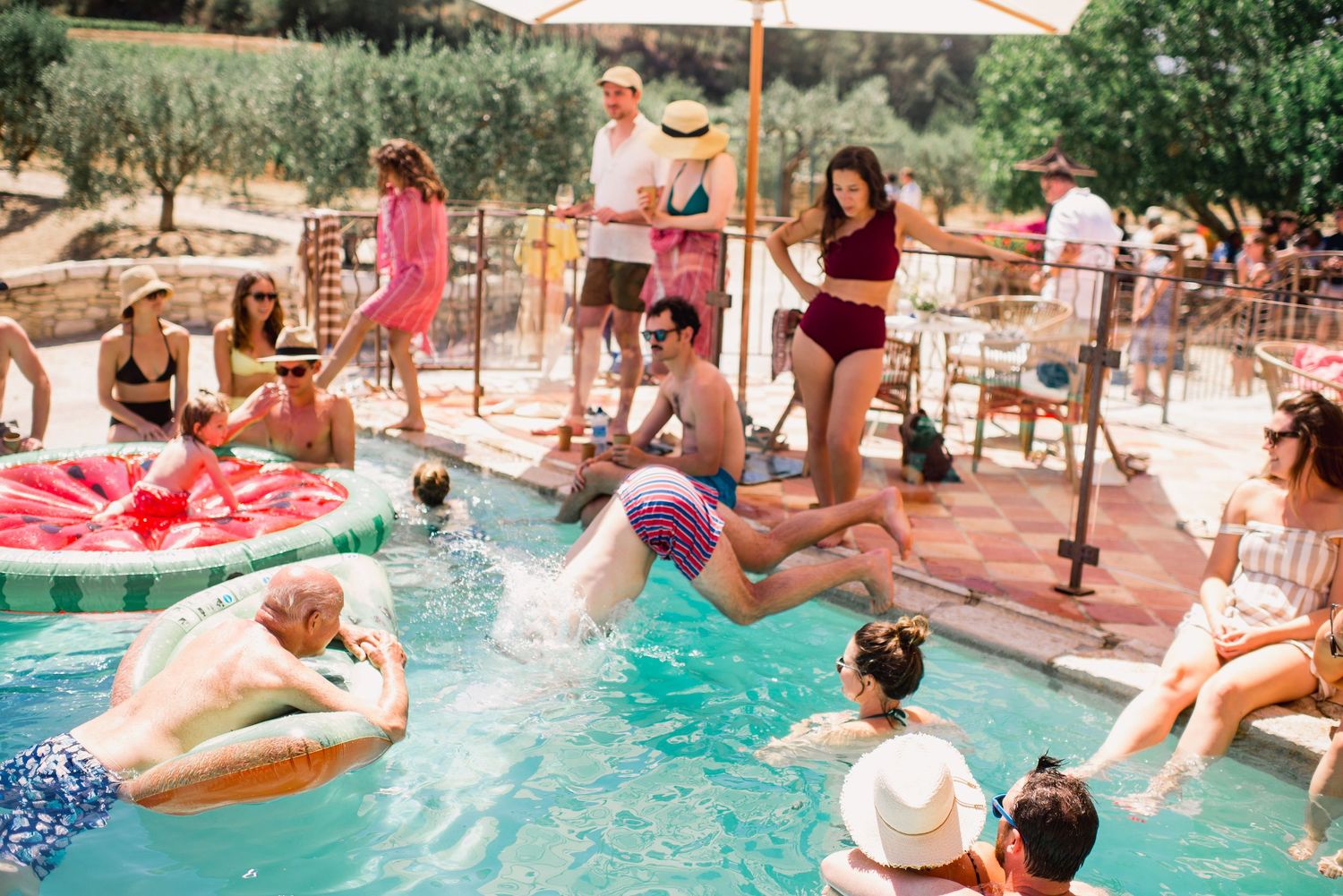 People enjoying a summer pool party with floaties and loungers at an outdoor venue surrounded by olive trees.