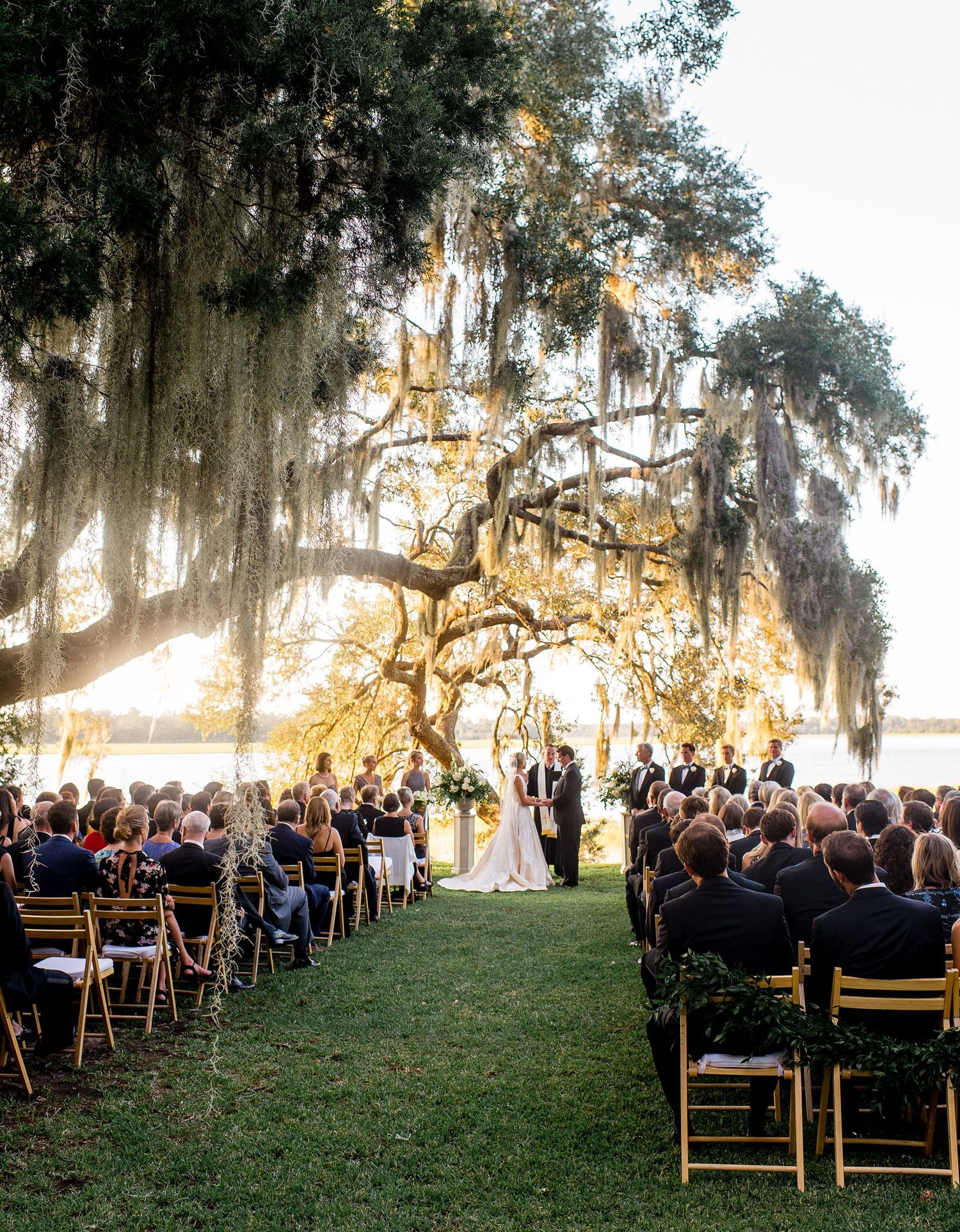 Outdoor wedding ceremony setup with wooden chairs under moss-draped oak trees at sunset.