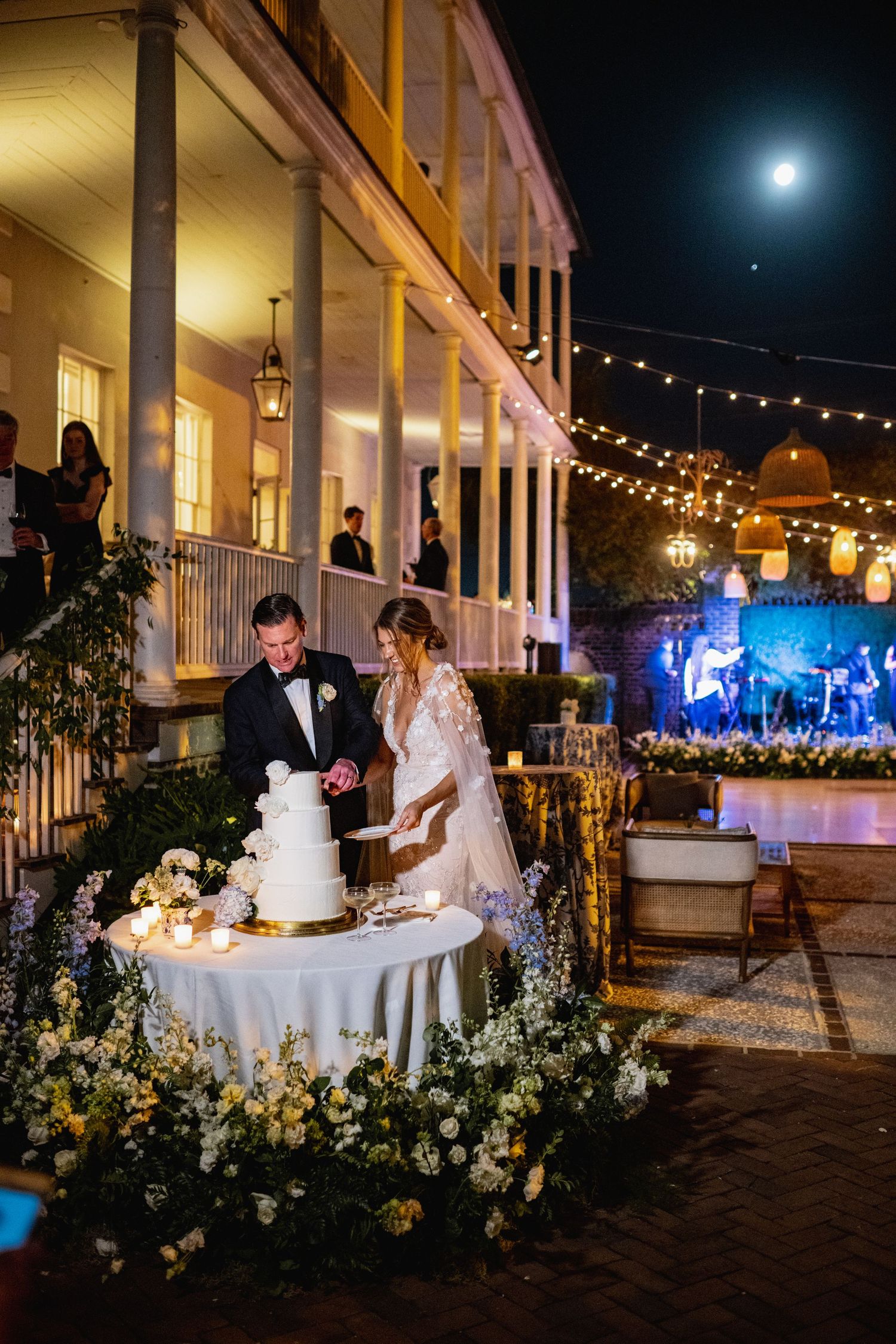 Romantic evening wedding reception under string lights with elegant table settings and floral arrangements on a patio.