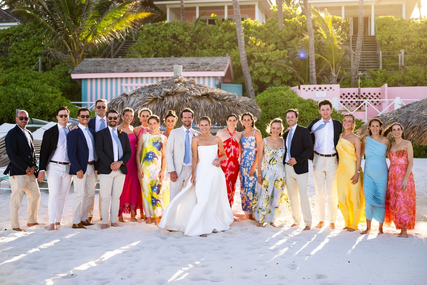 A large wedding party poses outdoors in formal attire with bridesmaids in colorful dresses and groomsmen in suits.