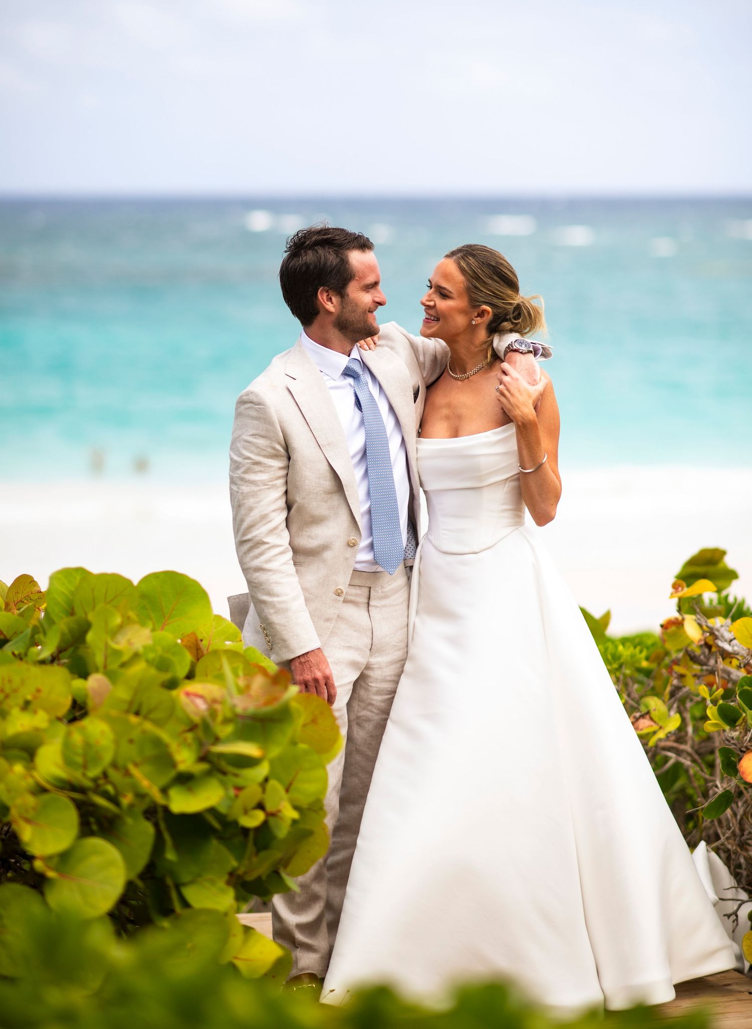 A romantic beach wedding couple poses by tropical foliage with turquoise ocean waters in the background.