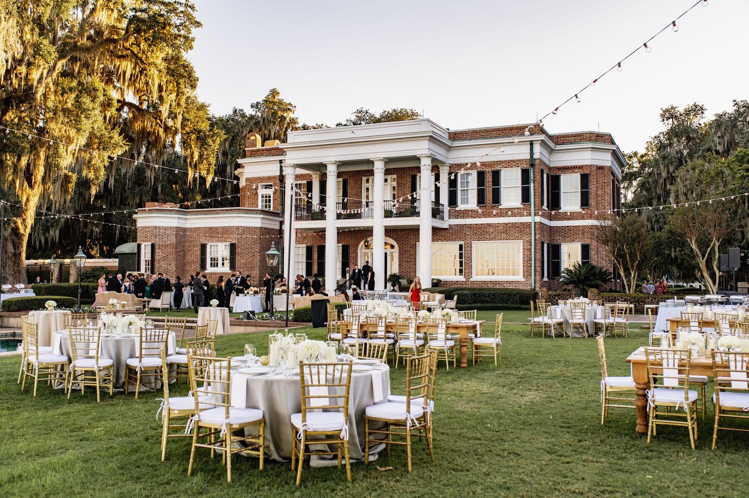 An elegant outdoor wedding reception setup with white chairs and round tables on a manicured lawn near a historic mansion.