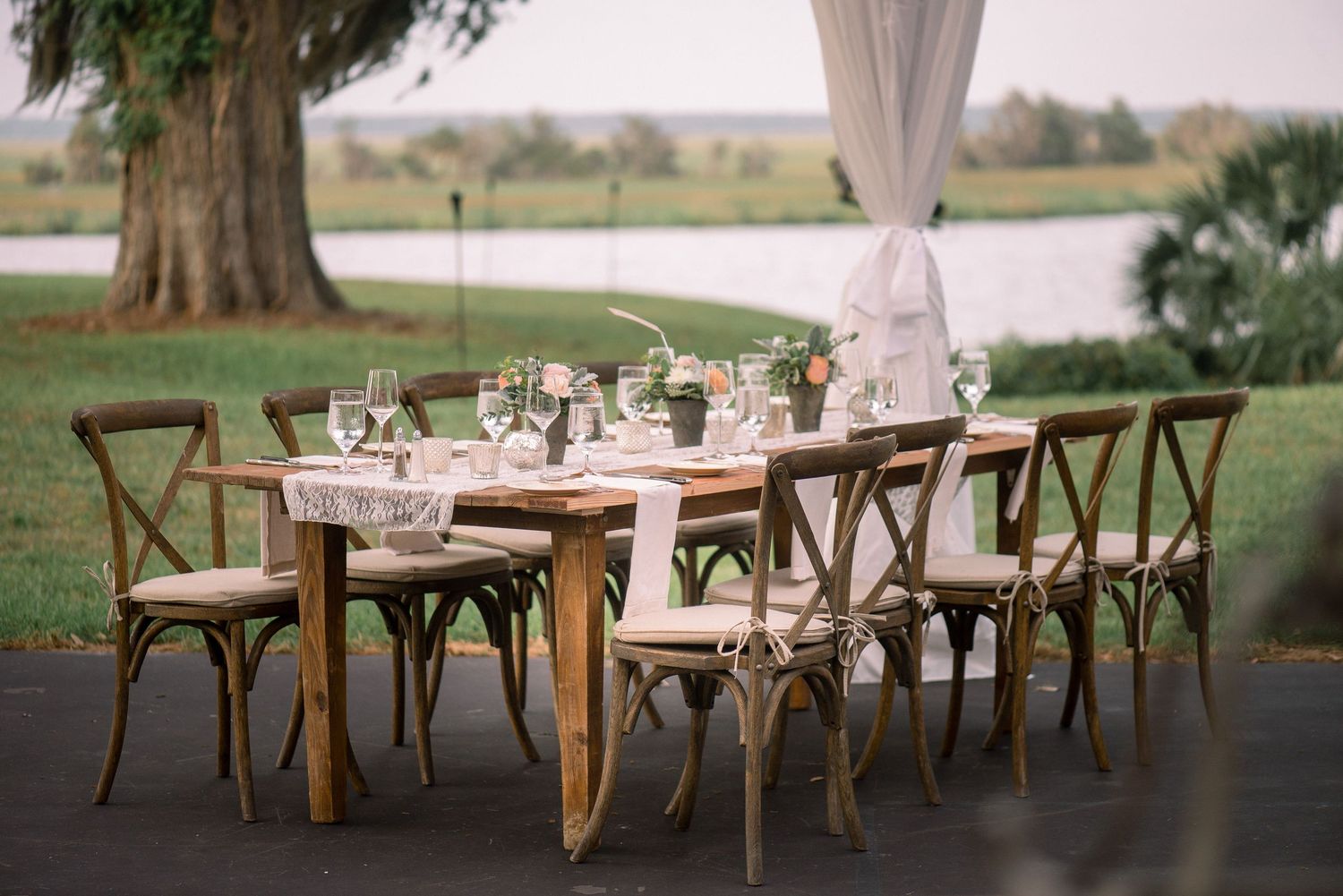 Long rustic wooden tables with crossback chairs set up outdoors under a white canopy for an elegant garden reception.
