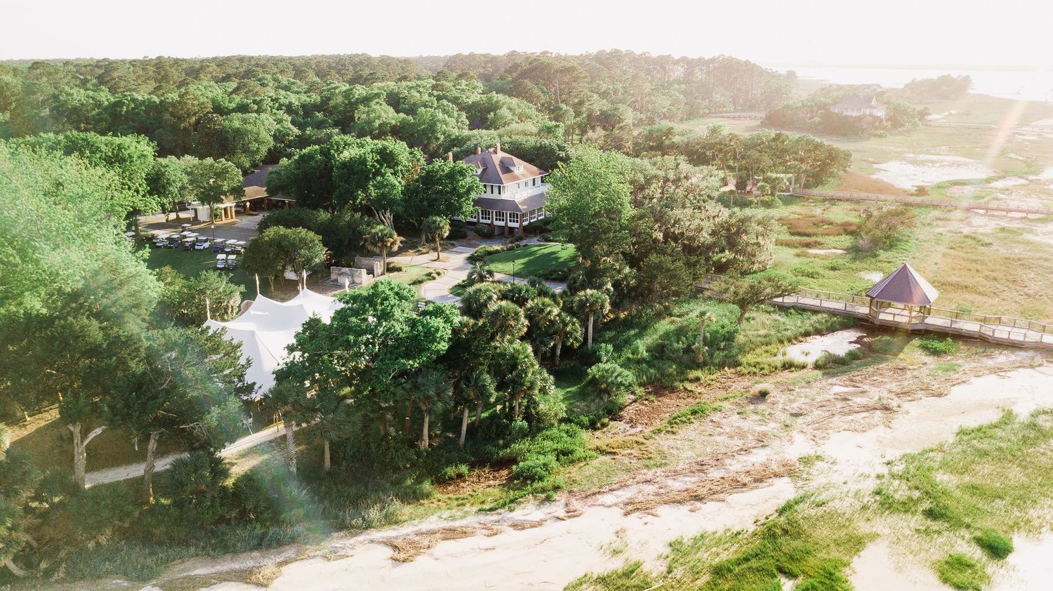 Aerial view of a coastal landscape with pine trees and scattered buildings along a sandy beach at sunset.