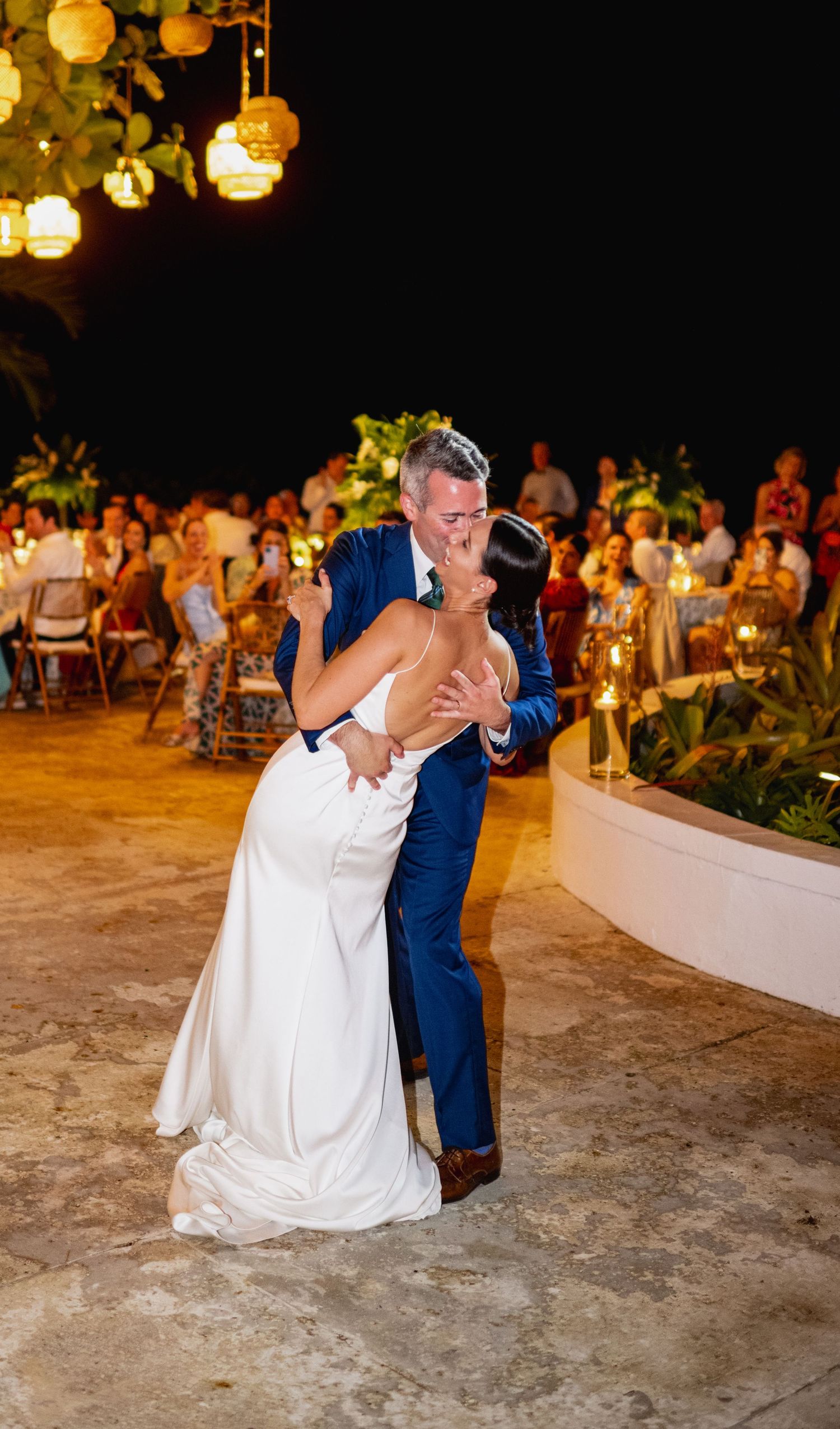 A couple shares their first dance at an evening wedding reception with glowing lanterns and guests in the background.