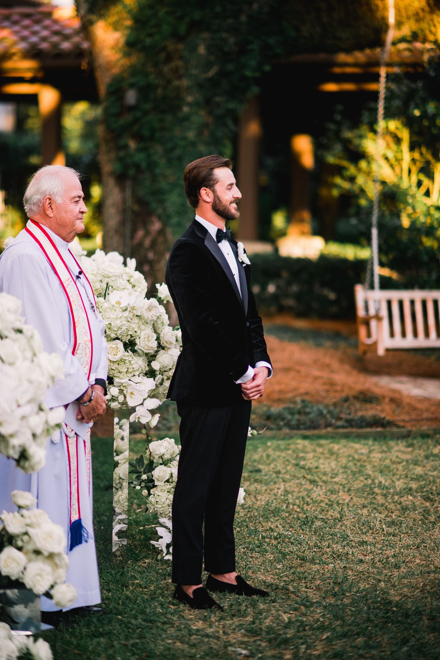A wedding ceremony setup outdoors with white floral arrangements and a clergyman in traditional robes.