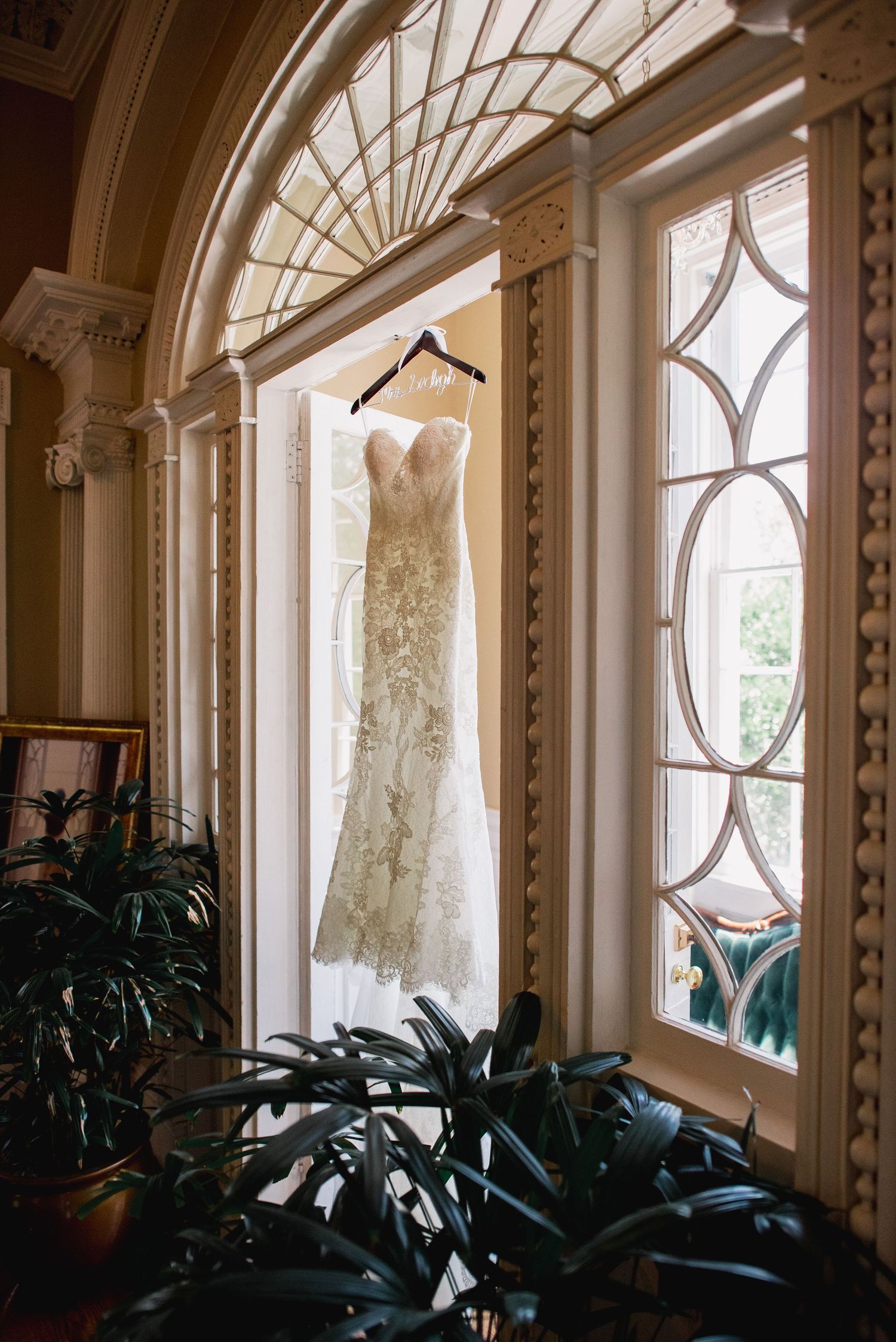 A white lace wedding dress hangs elegantly in front of ornate windows with decorative trim and indoor plants.