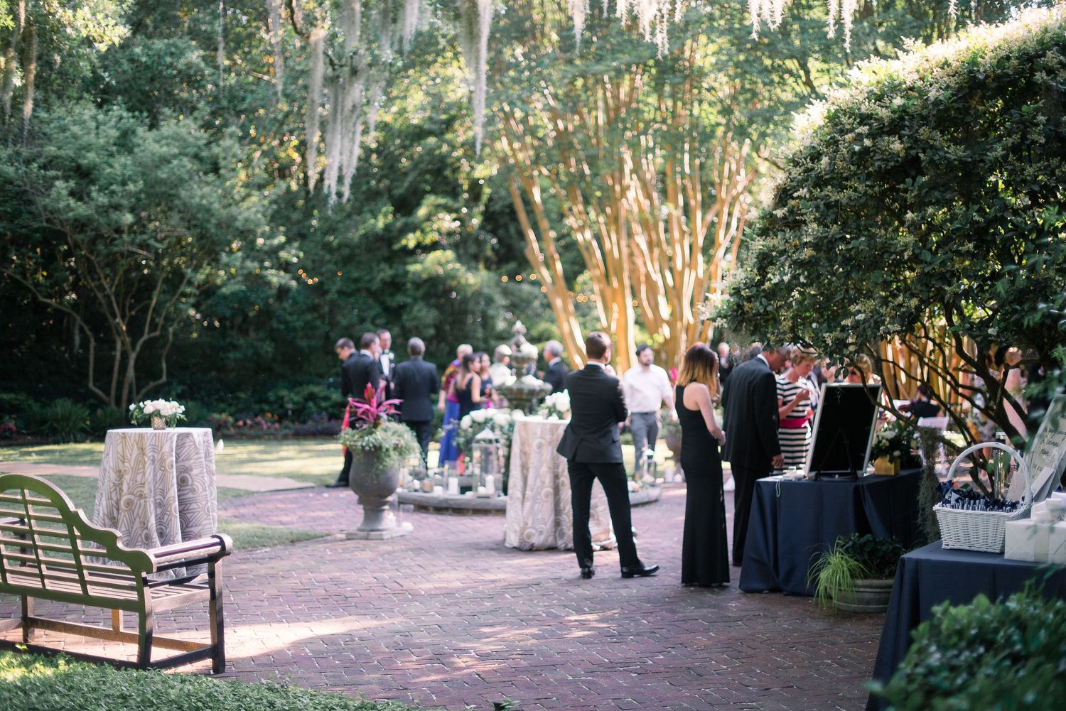 Outdoor garden party reception with guests mingling around cocktail tables and buffet stations in a lush wooded setting.