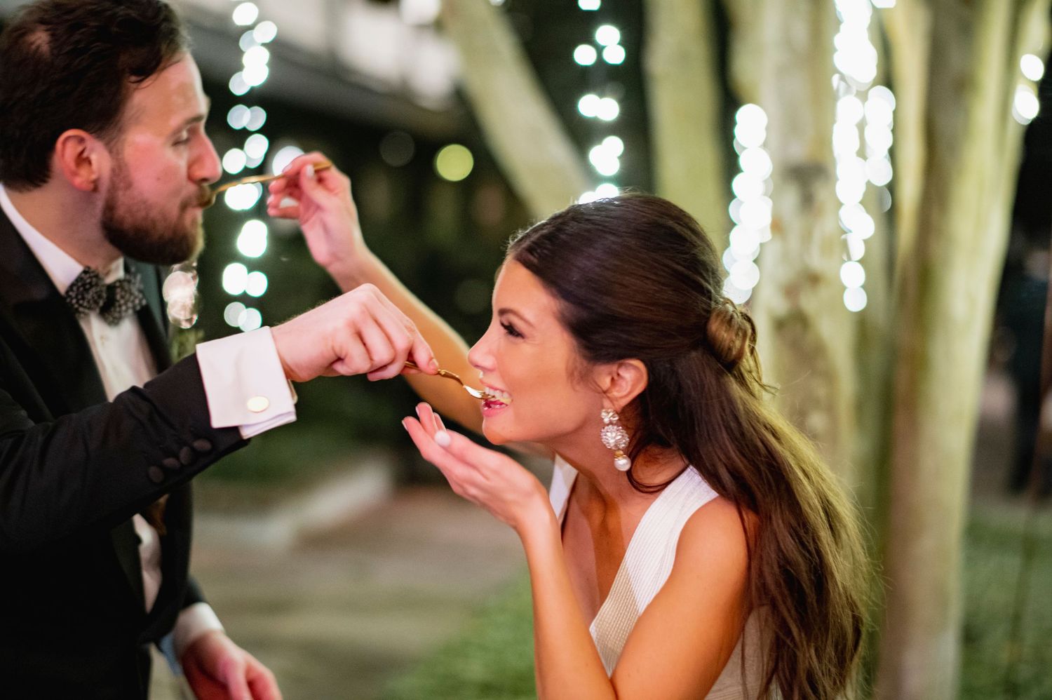 A romantic wedding couple shares an intimate moment under twinkling lights in a tree-lined outdoor setting.