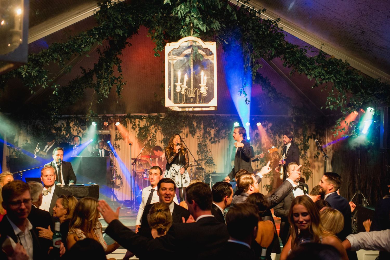 A lively wedding reception dance party with colorful lighting and greenery decorations in a rustic venue.
