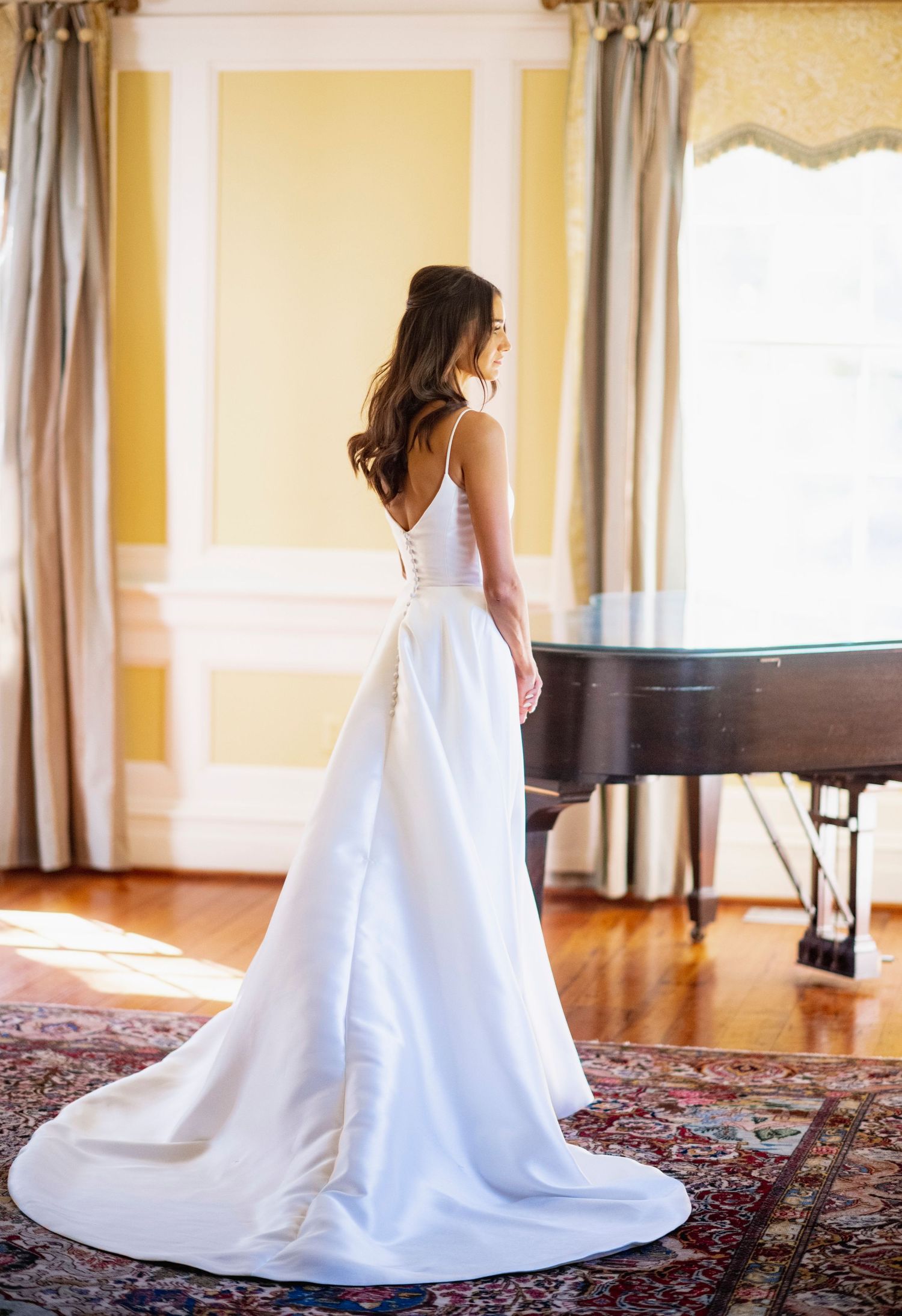 Elegant white wedding gown with train photographed in a sunlit room with yellow walls and a grand piano.