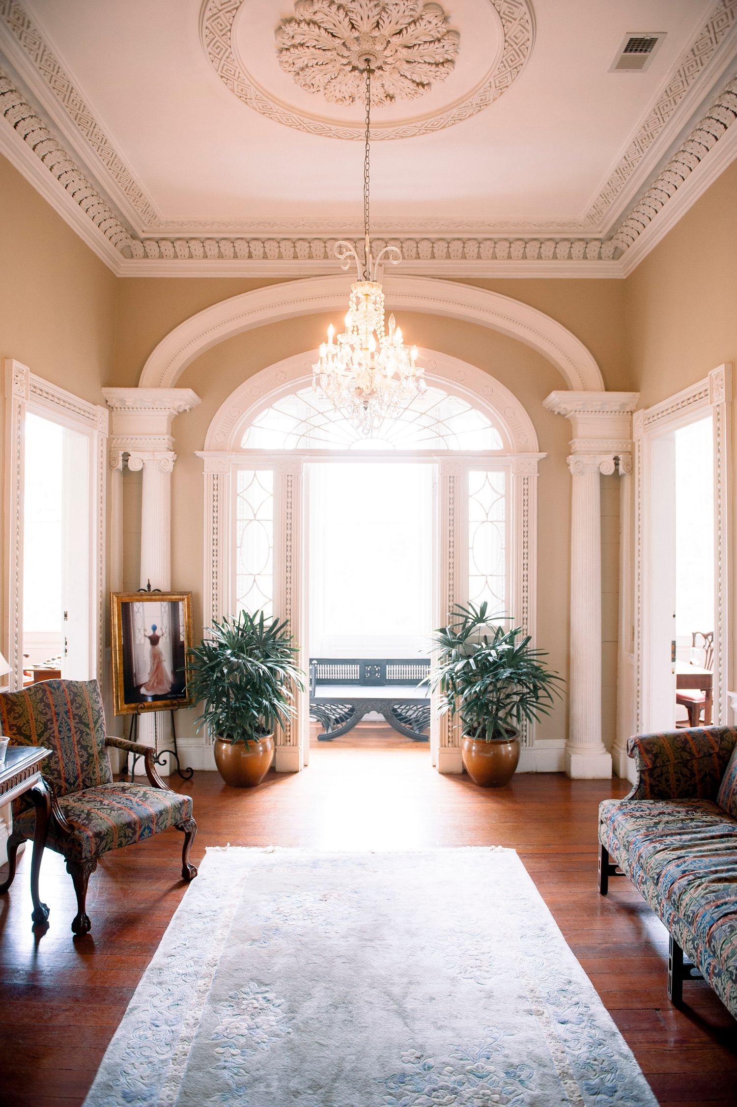 Elegant white ballroom with ornate crown molding, crystal chandeliers, hardwood floors and arched windows in panoramic view.