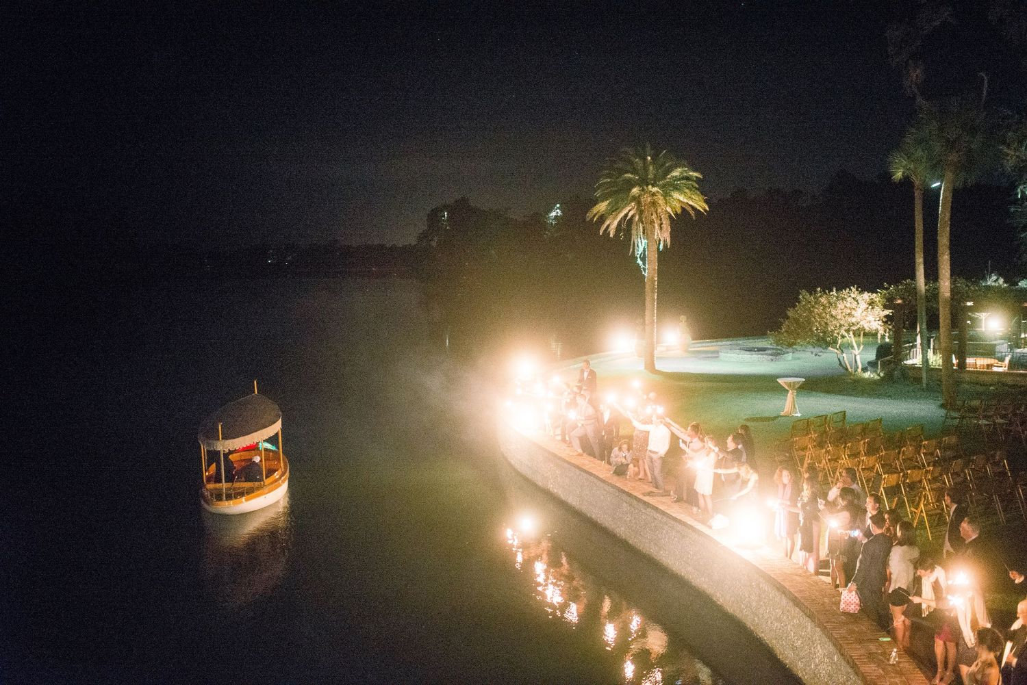 Nighttime wedding ceremony along illuminated waterfront with boat and palm trees creating romantic ambiance.