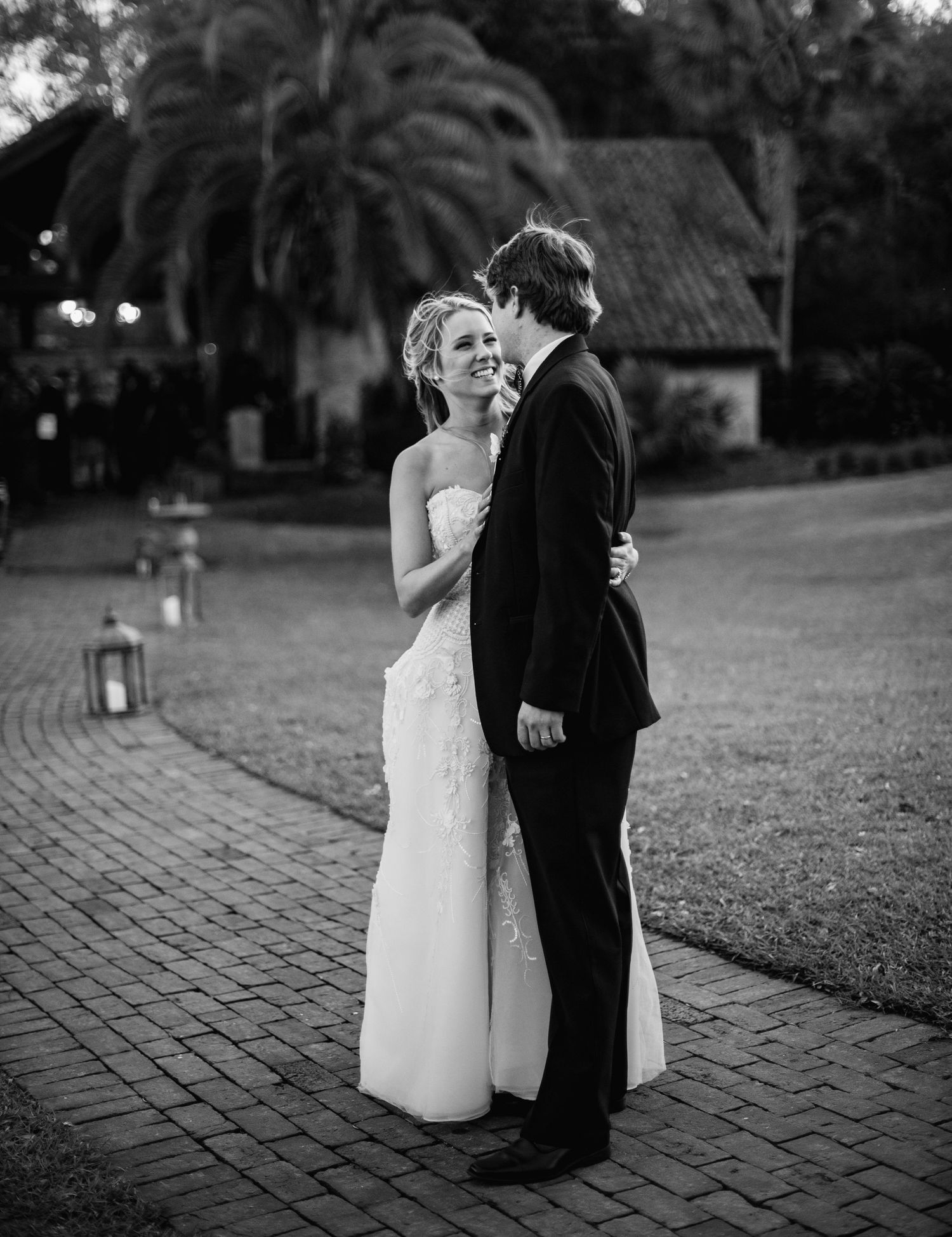 A couple shares a romantic moment during their wedding on a brick pathway with palm trees and lanterns in the background.