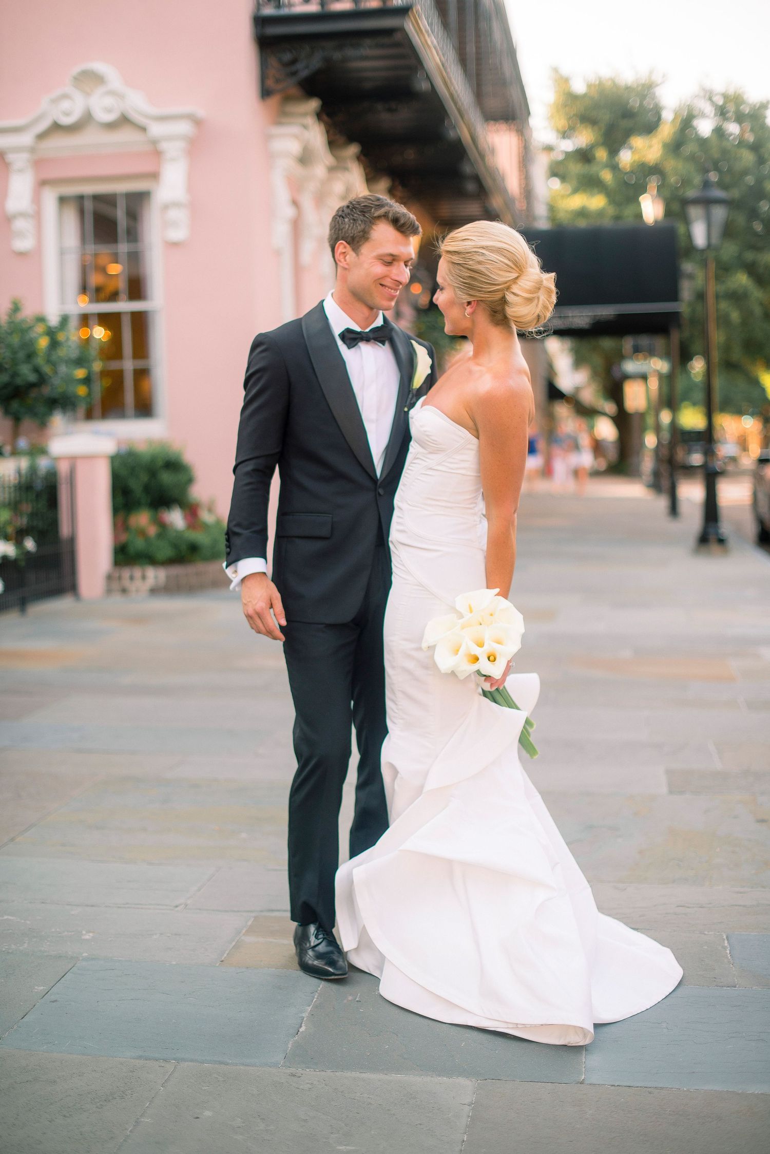 A romantic wedding couple in formal attire dance together on a paved courtyard near a pink building at sunset.