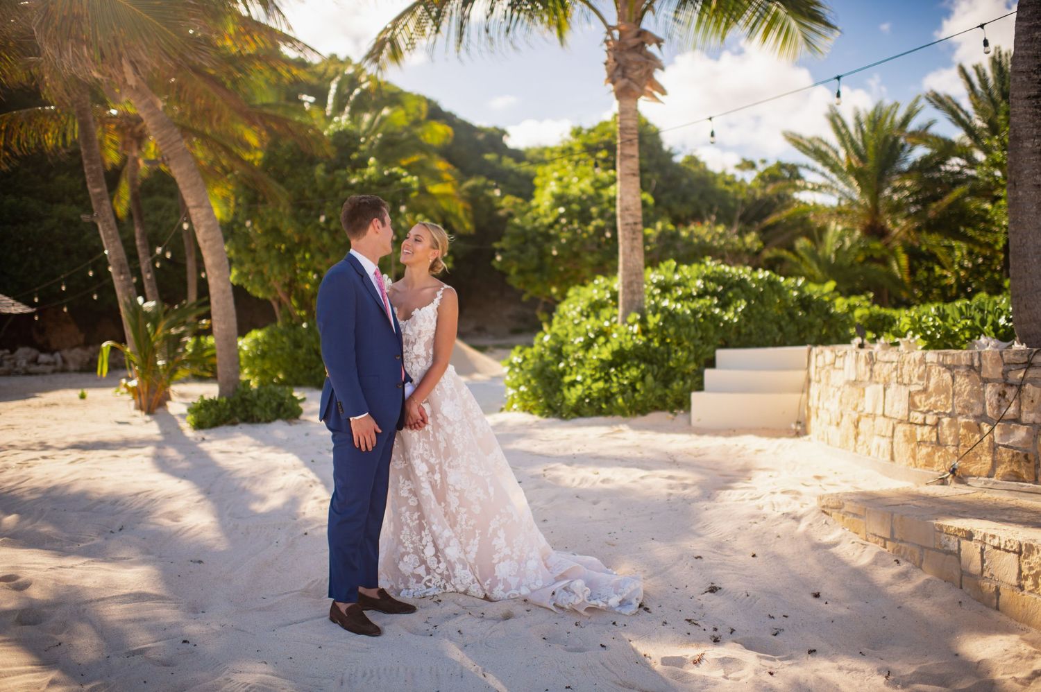 Newlyweds share romantic moment on tropical path surrounded by palm trees and stone walls.