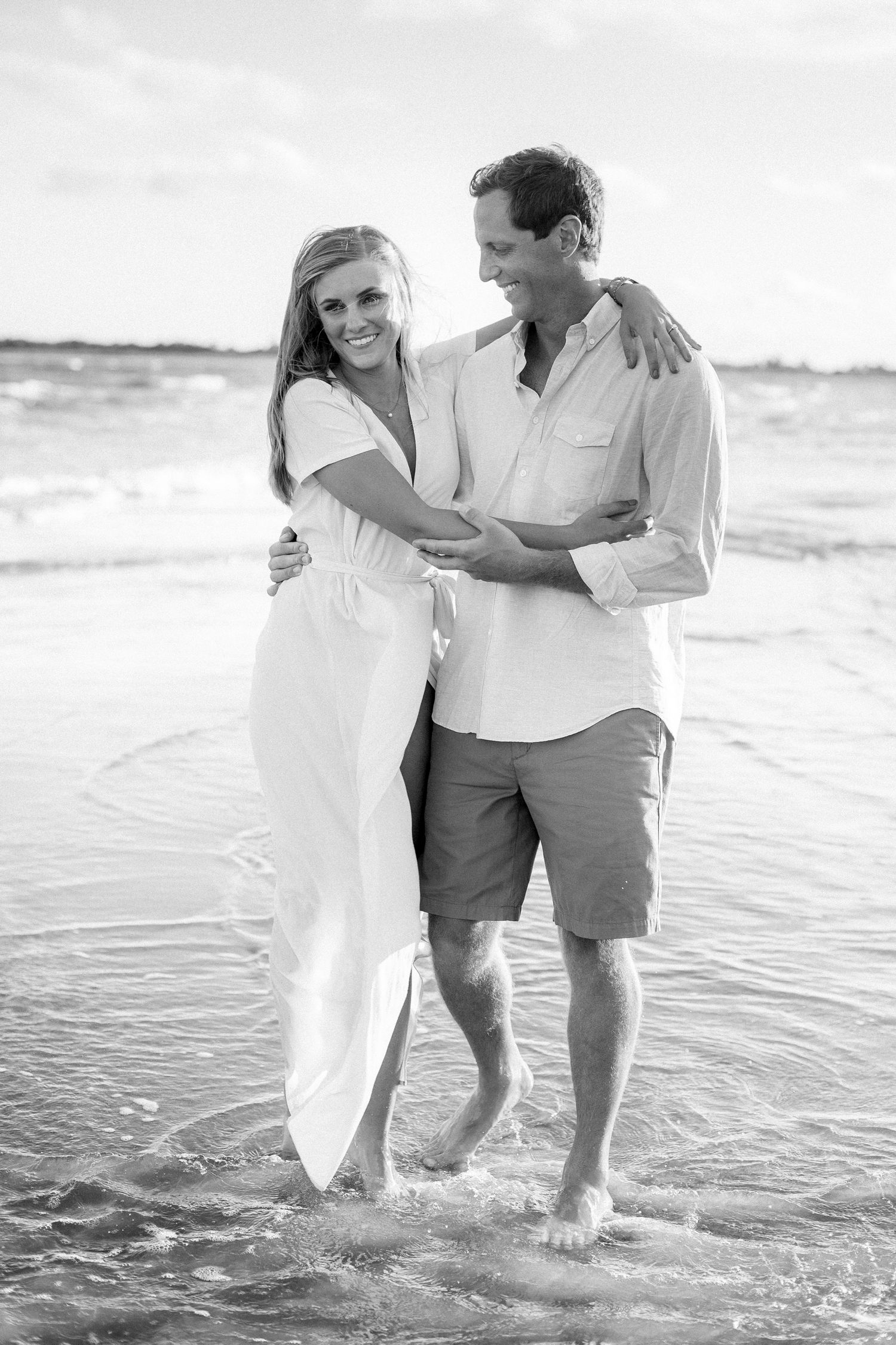 Romantic couple walking in shallow ocean waves wearing white and casual attire in black and white photo.