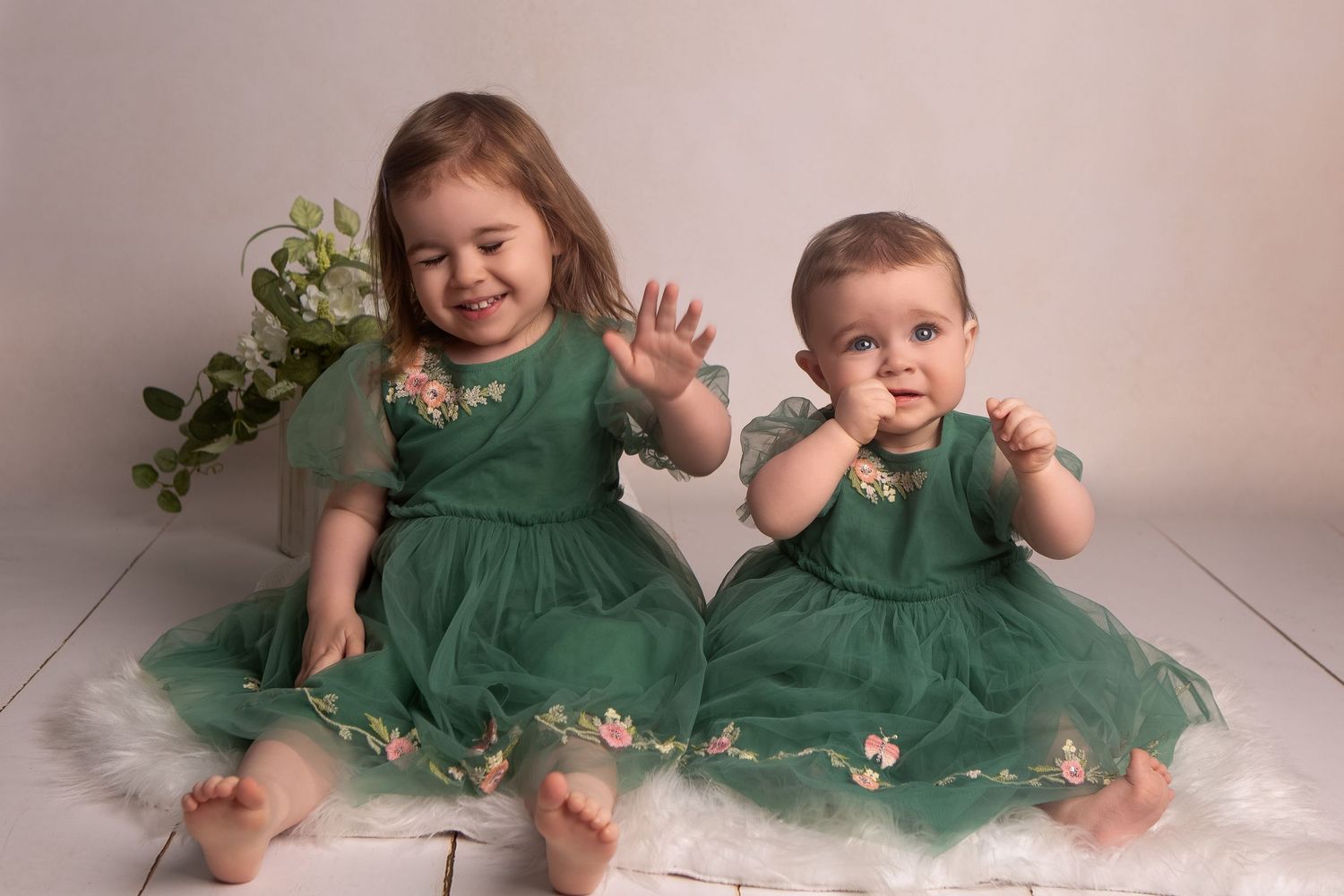 Two baby girls in matching green floral dresses sitting together on a white fur rug, smiling and waving.