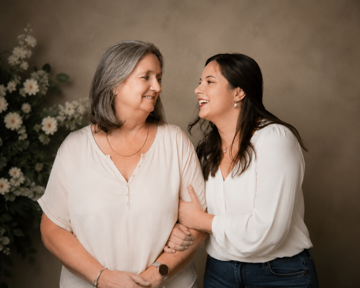 Two women smiling and embracing warmly in a professional studio portrait with floral backdrop.